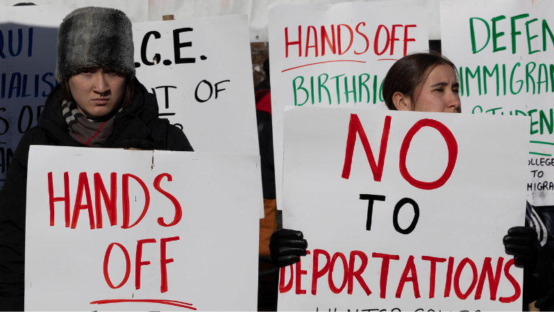 Das Foto zeigt zwei junge Frauen bei einer Demonstration, die Schilder hochhalten. Auf dem linke steht "Hands off", auf dem rechten "No to deportations".