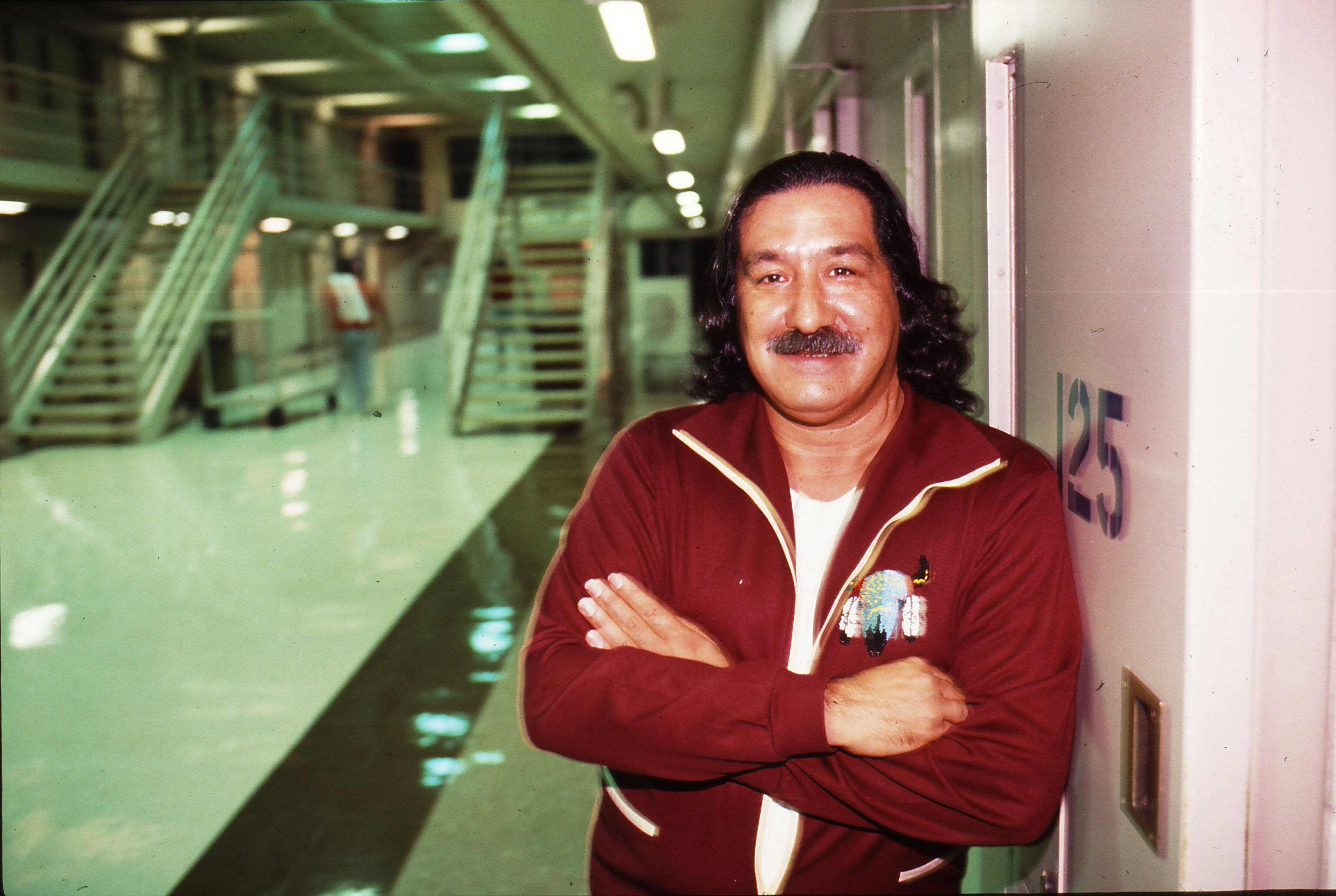Das Foto zeigt Leonard Peltier in einem Gefängnisflur. Er lehnt mit verschränkten Armen an der Wand und lächelt in die Kamera.