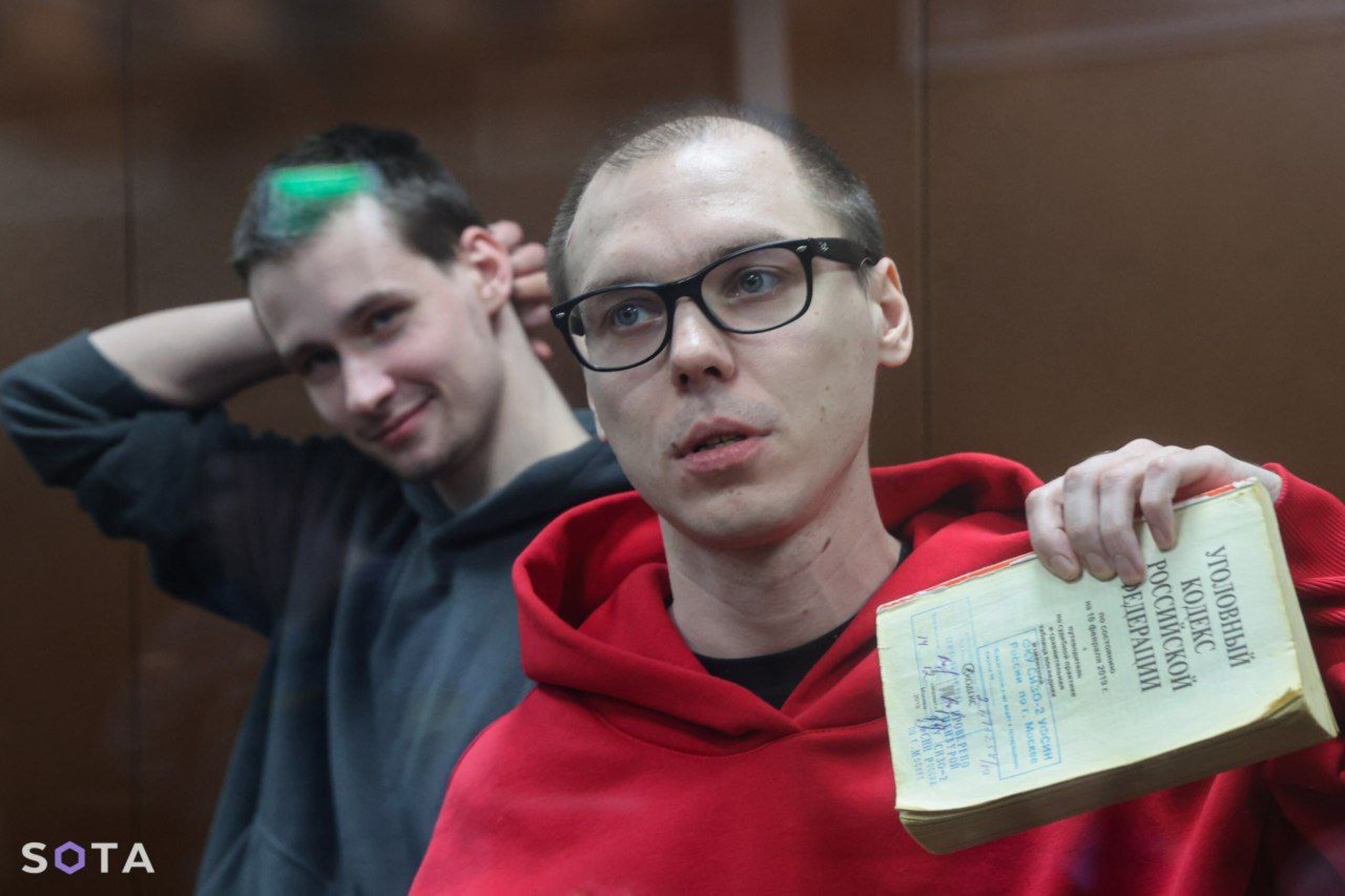 Das Foto zeigt Artyom Kamardin. Er trägt eine Brille und hält mit seiner linken Hand ein aufgeschlagenes Buch hoch. Yegor Shtovb steht hinter ihm und lächelt und kratzt sich mit der rechten Hand am Hinterkopf.