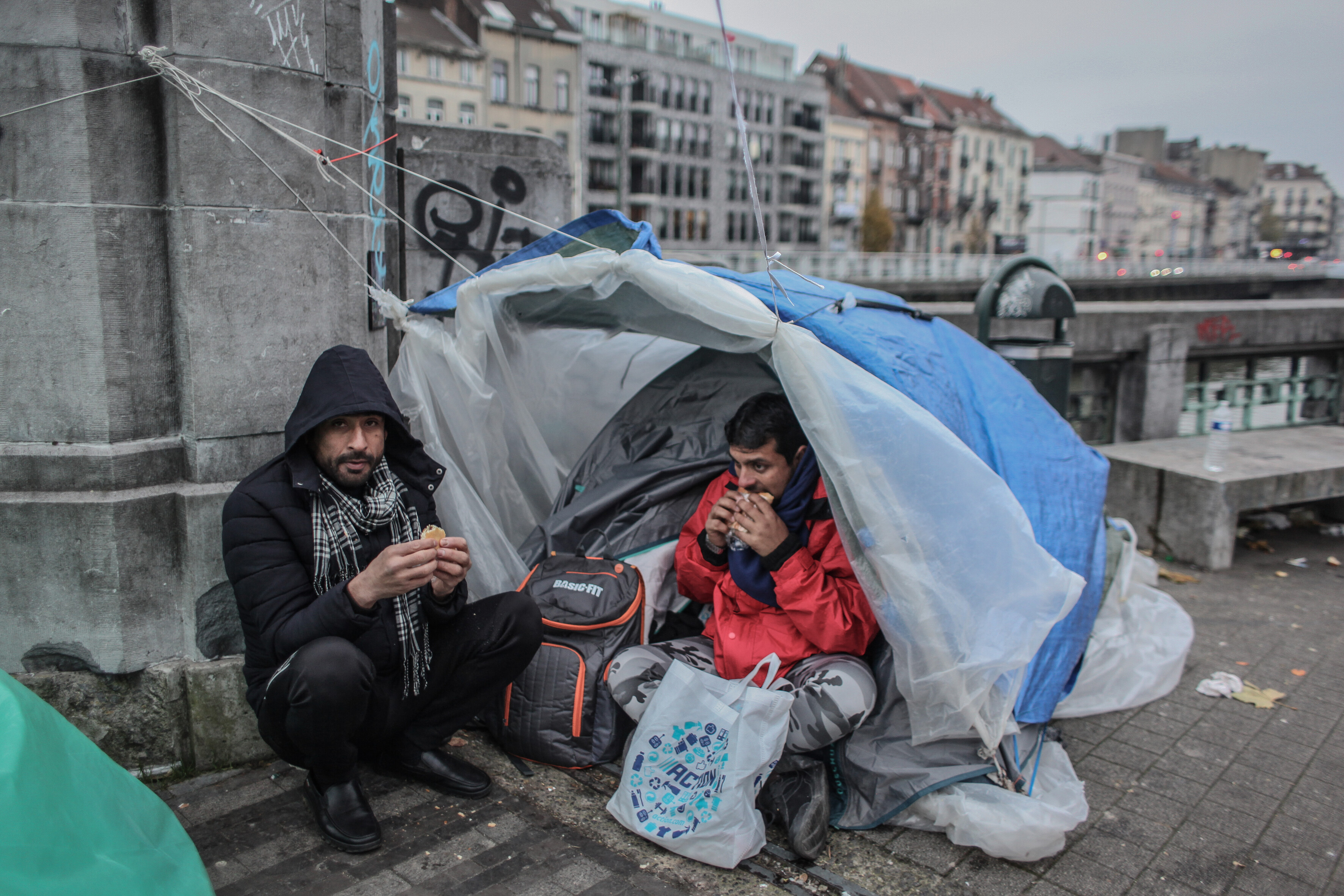 Das Foto zeigt zwei Männer vor einem Campingzelt kauernd, das auf einem Bürgersteigt errichtet wurde. Sie essen etwas.