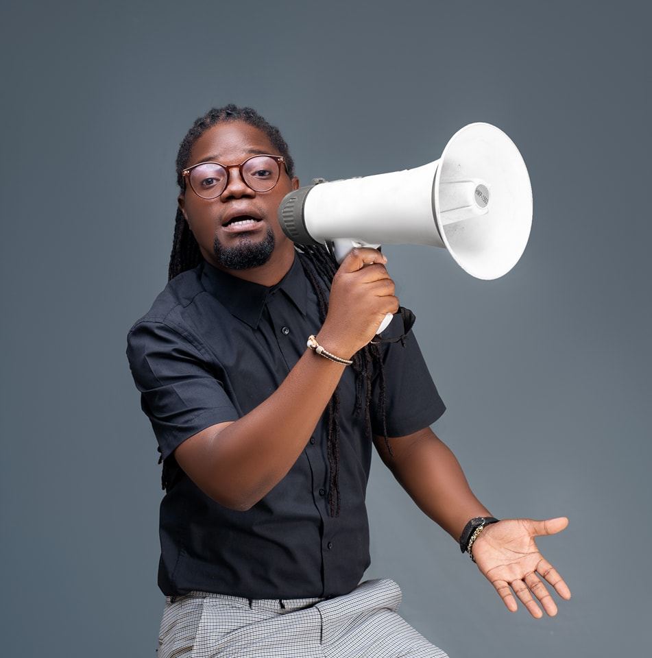 Das Foto zeigt Joel Amaral bei einem Fotoshooting vor neutralem Hintergrund. Er trägt eine Brille und hält mit seiner rechten Hand ein Megafon vor seinen Mund.