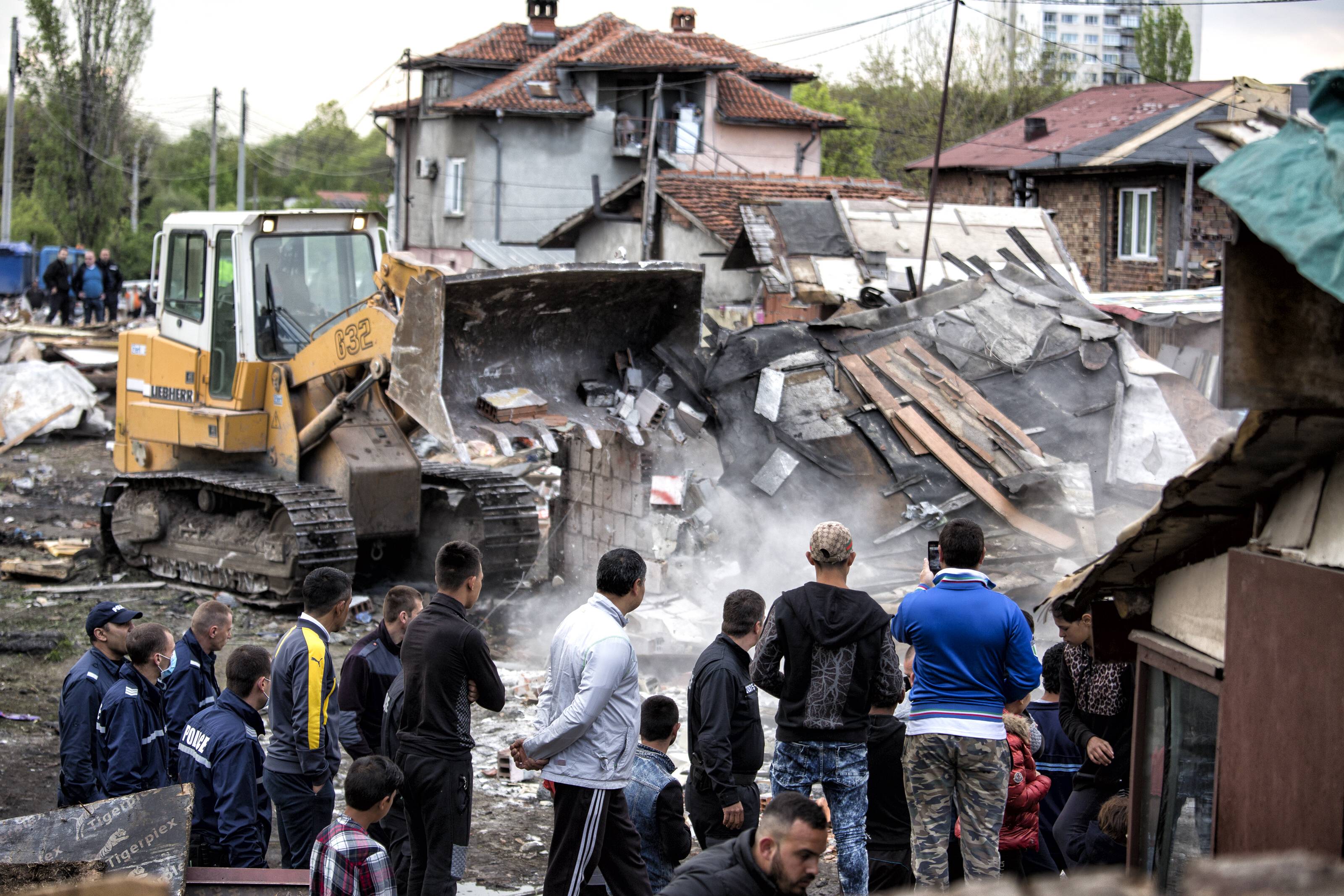Mehrere Personen beobachten, wie ein Bulldozer über die Trümmer zerstörter Häuser fährt.