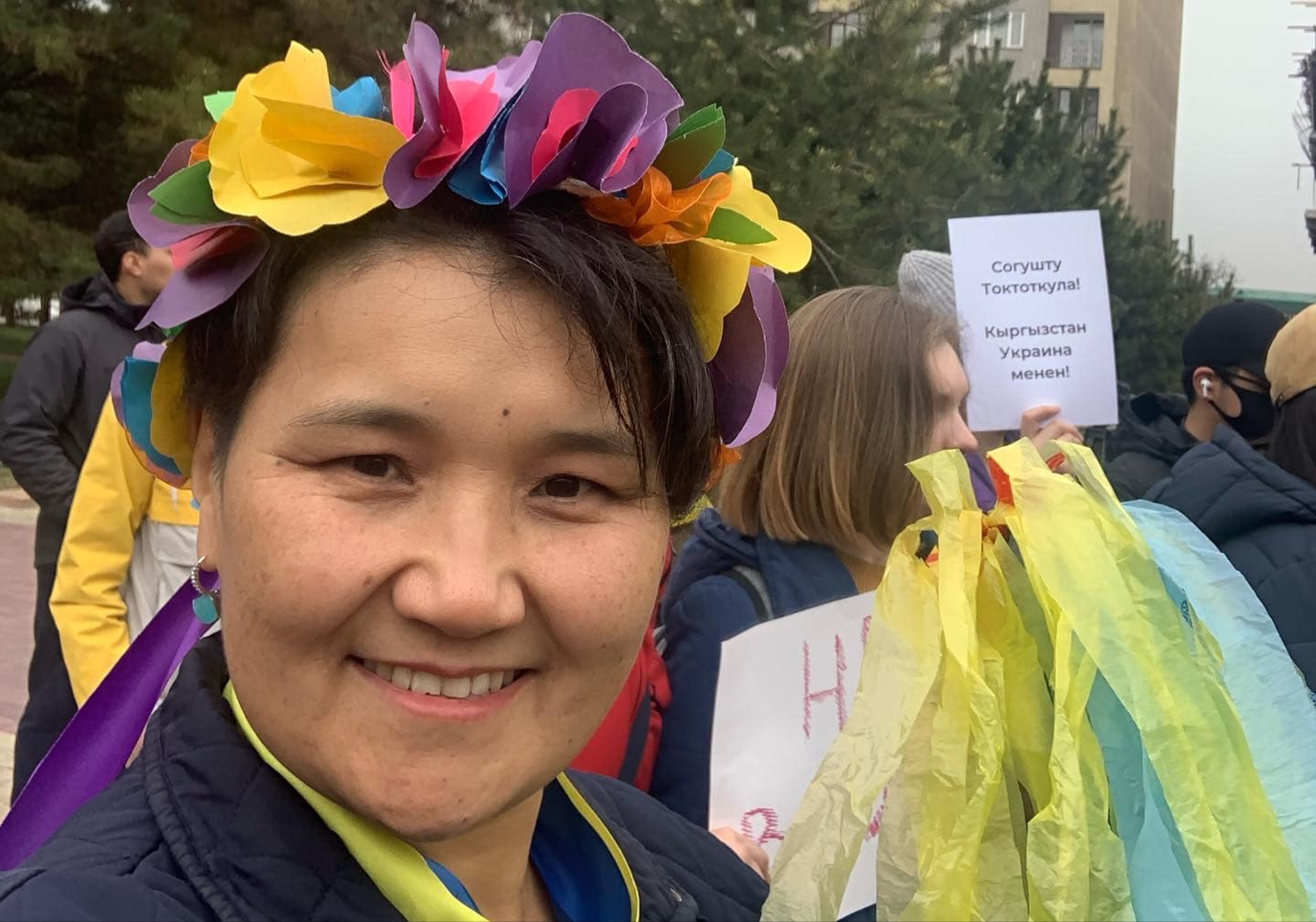 Das Foto zeigt Rita Karasartova bei einer Protestaktion. Sie trägt einen Haarkranz mit vielen verschiedenen Farben und lächelt in die Kamera.