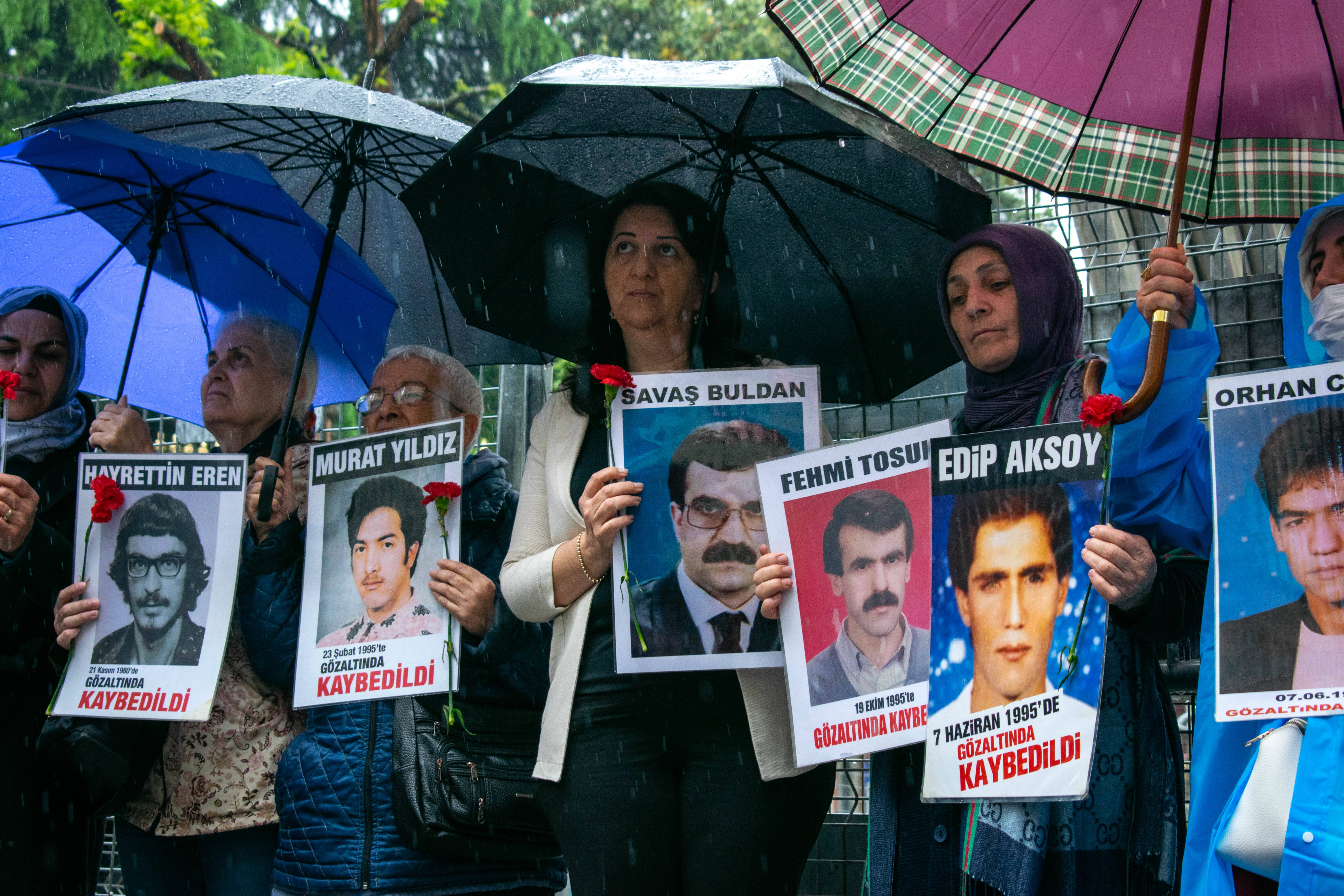 Das Bild zeigt mehrere Frauen mit aufgespannten Regenschirmen und Plakaten und Rosen in der Hand. Auf den Plakaten sind Fotos von Personen abgedruckt.