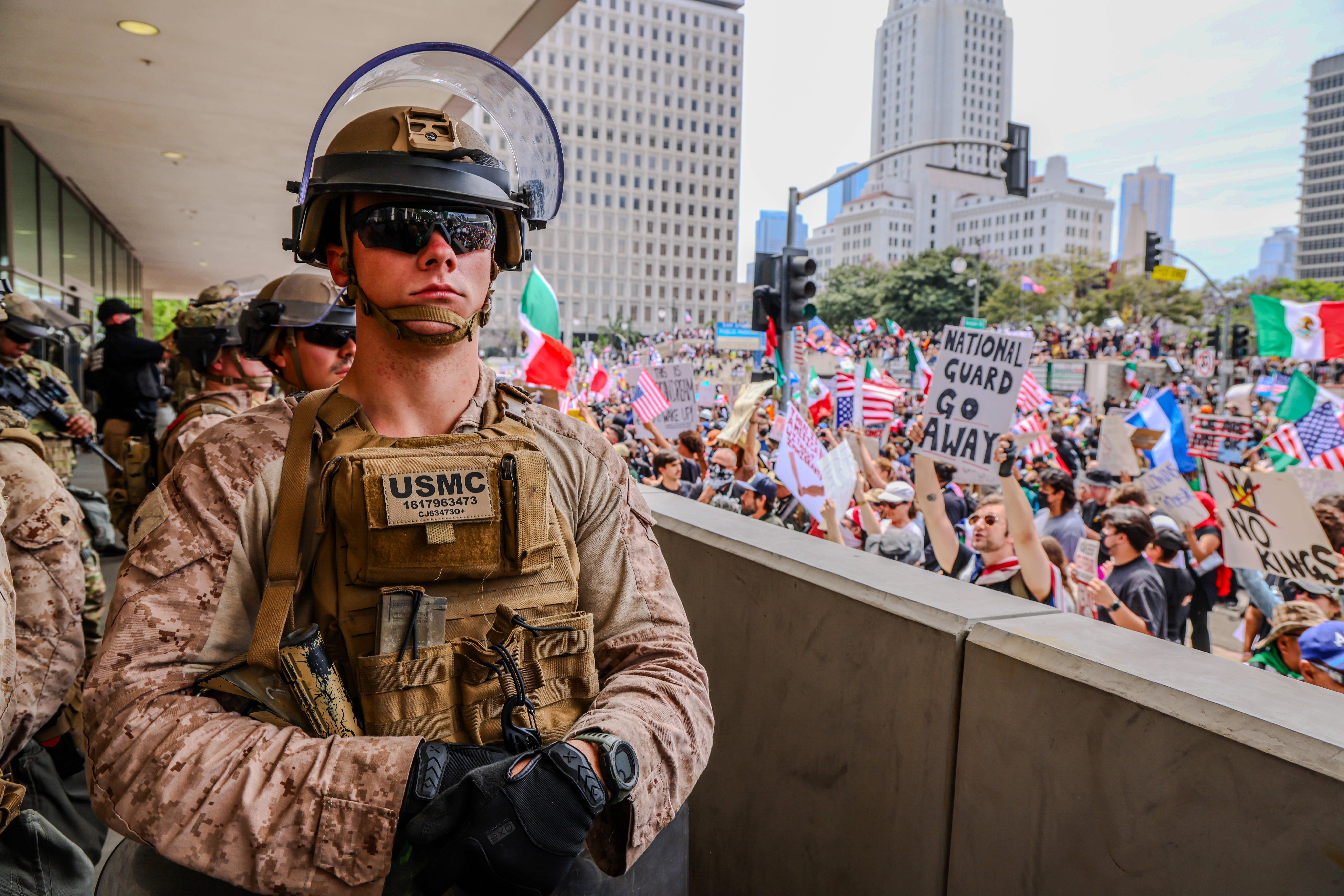 Das Foto zeigt einen Soldat mit Helm, Sonnenbrille und Schutzweste. Im Hintergrund demonstriert eine große Menschenmenge mit Fahnen und Bannern und Plakaten.