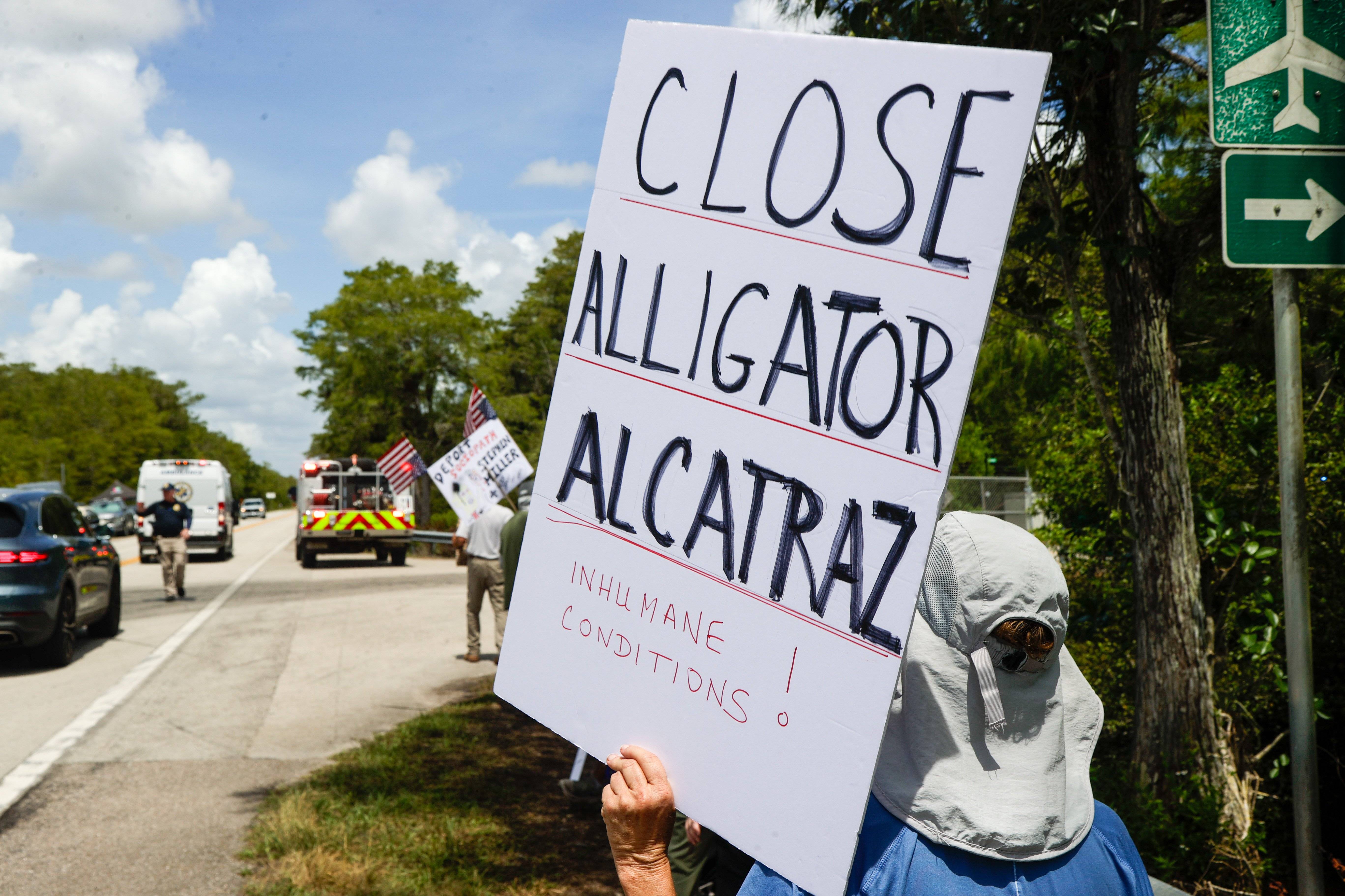 Das Foto zeigt eine Person am Straßenrand vor Bäumen. Sie hält ein Schild hoch, auf dem unter anderem steht: "Close Alligator Alcatraz".