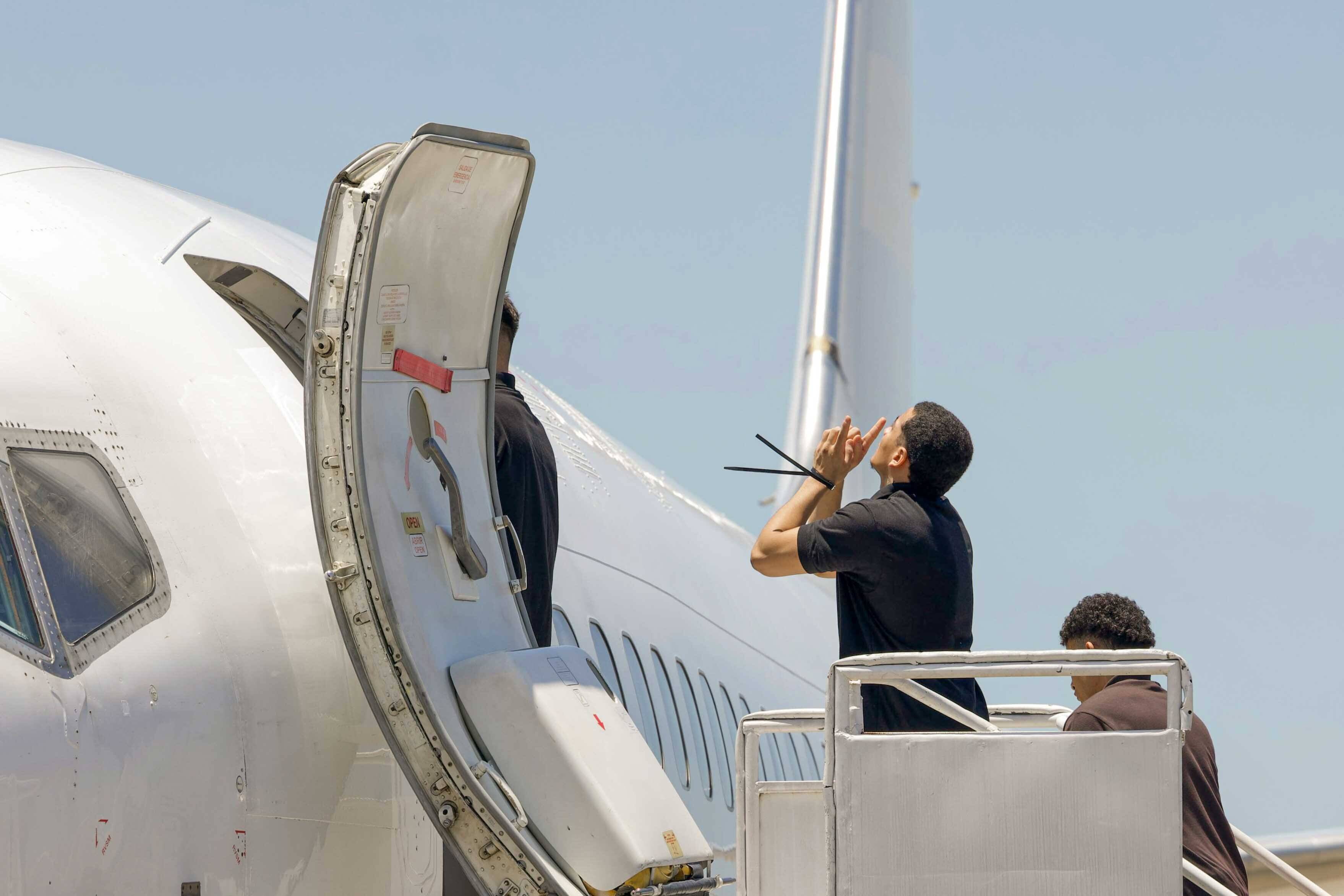 Das Foto zeigt mehrere Männer mit gefesselten Händen, die über eine Treppe in ein Passagierflugzeug einsteigen. Einer von ihnen hebt die Hände dankend Richtung Himmel.