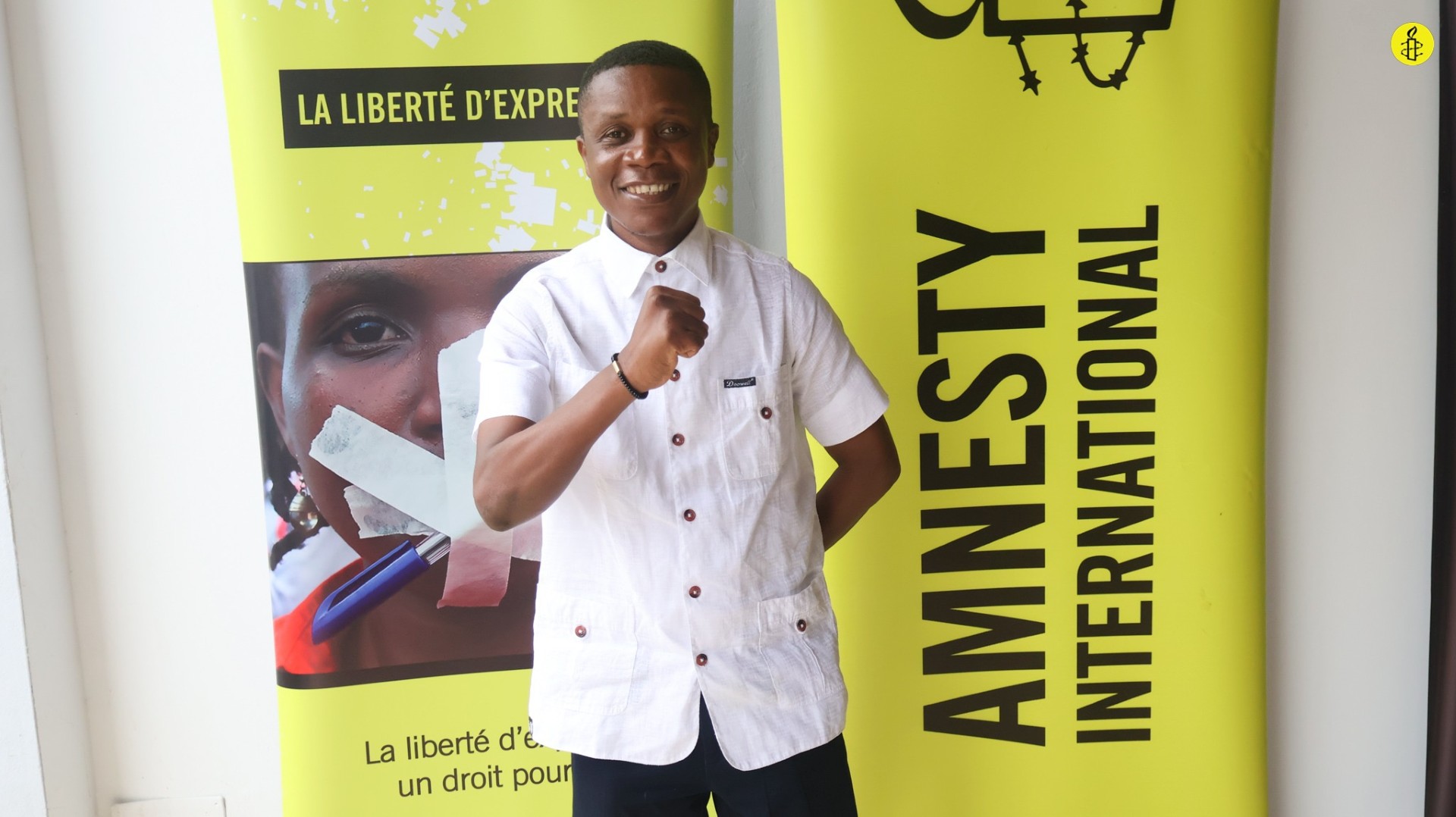 Ghislain Duggary Assy steht mit einem großen Lächeln vor einer Wand, an der unter anderem ein Banner angebracht ist mit dem Amnesty-Logo und dem Schriftzug "Amnesry International". Er ballt die rechte Faust und hält sie vor sich.