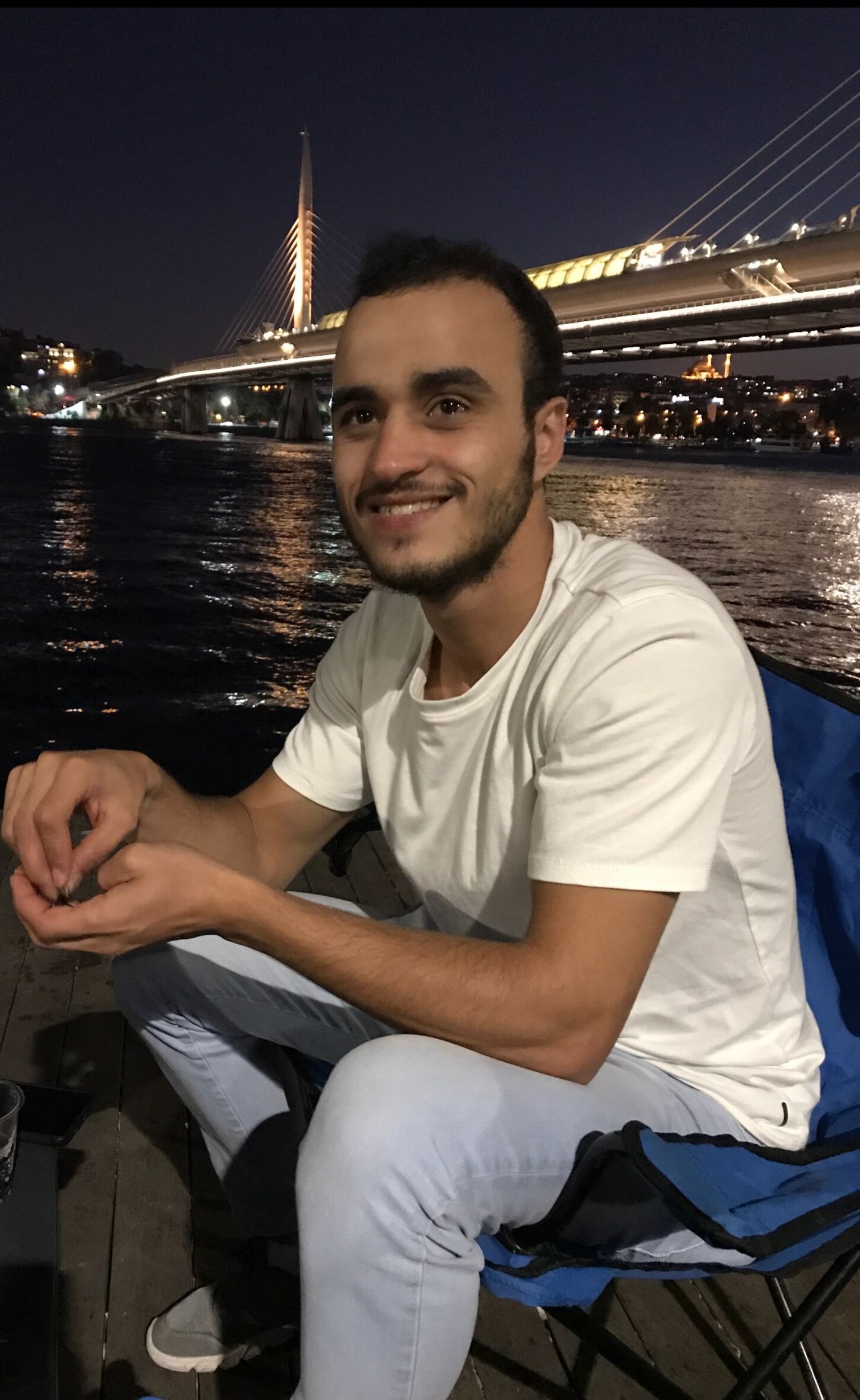 Foto von Ahmad Aabo, der nachts an einem Gewässer sitzt, in dem sich die Lichter der Stadt spiegeln. Er schaut an der Kamera vorbei und lächelt.