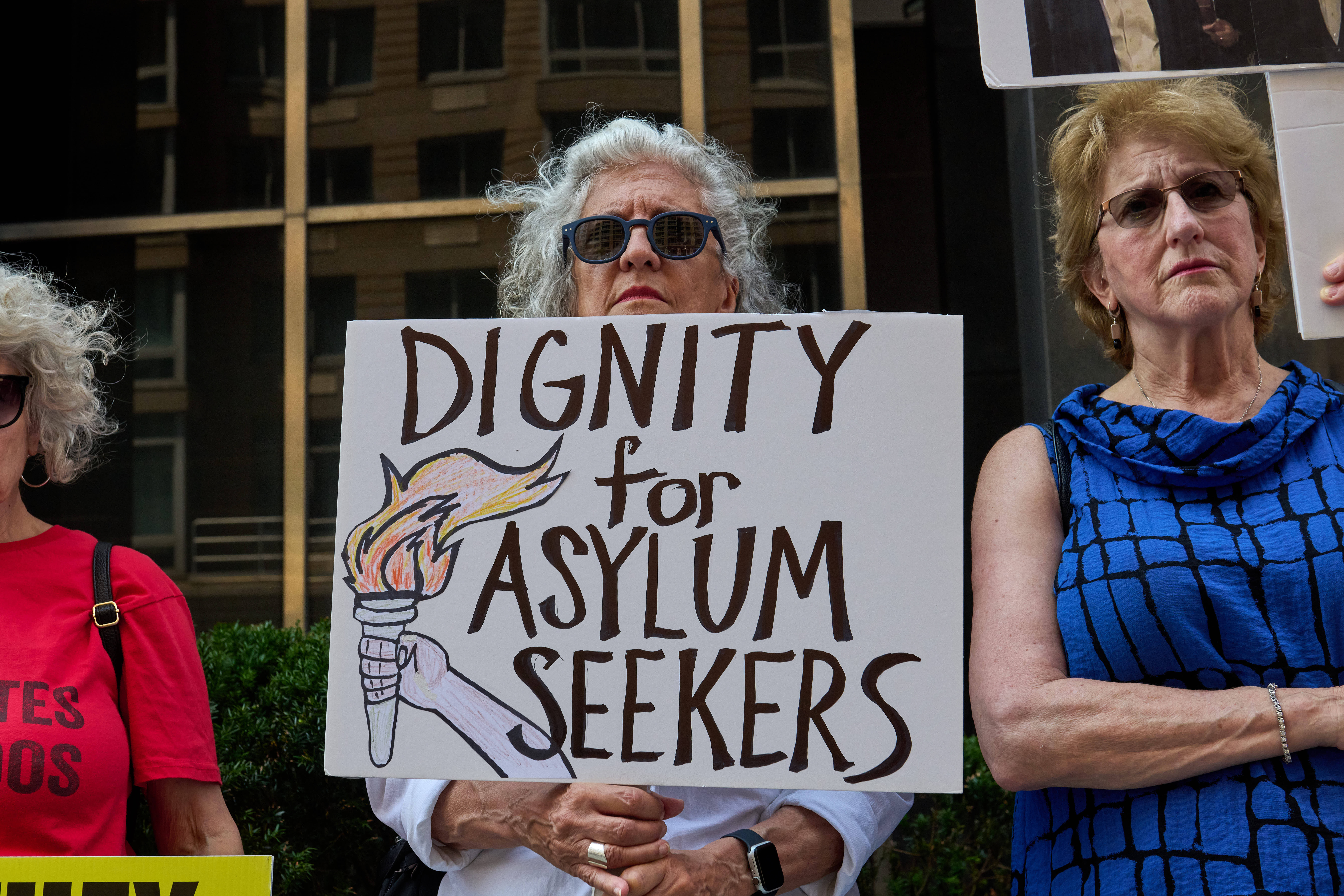 Das Foto zeigt mehrere Frauen bei einer Protestaktion vor einem Gebäude. Die Frau in der Mitte hält ein Schild hoch, auf dem steht: "Dignity for Asylum Seekers".