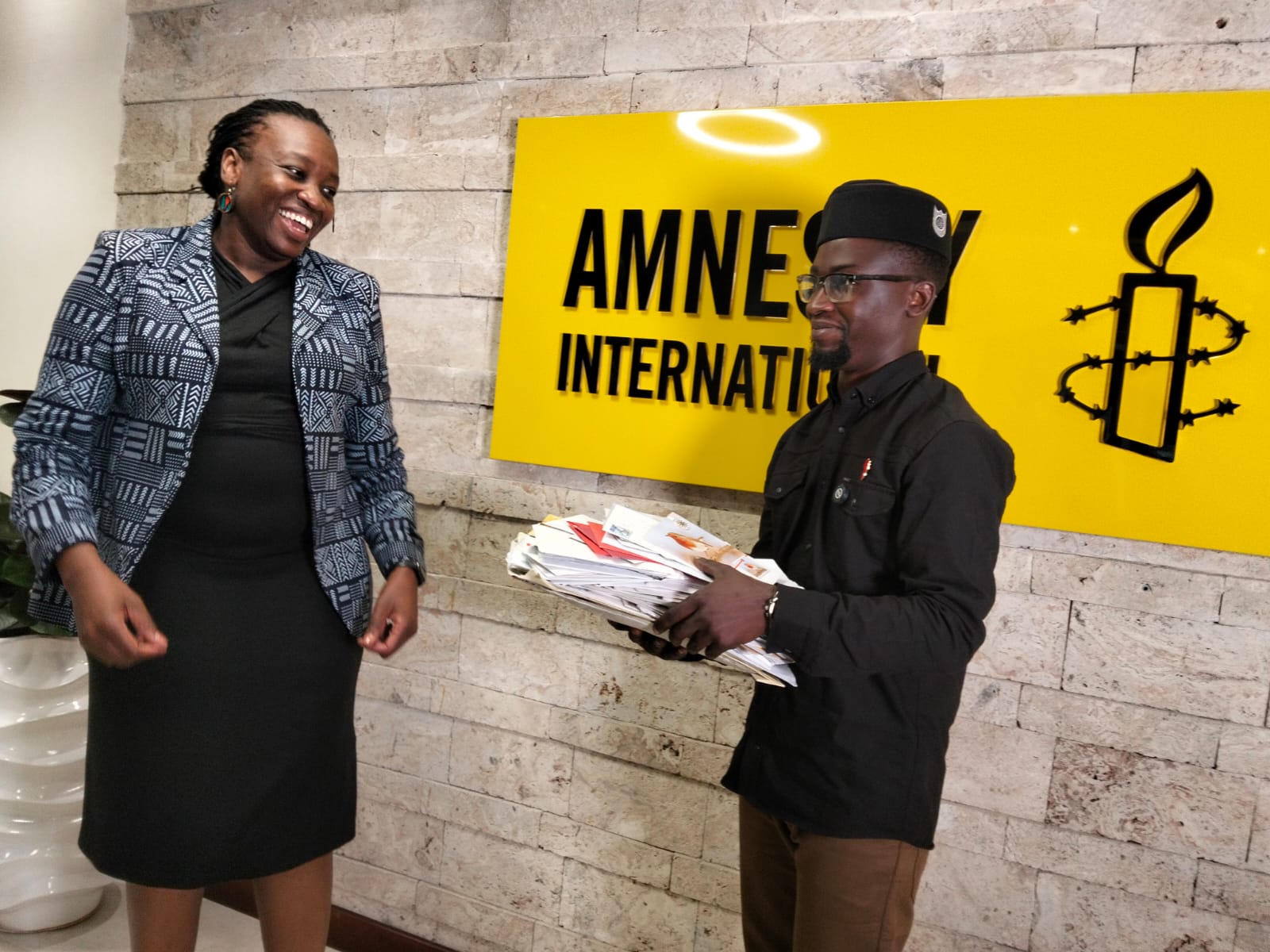 King Mwamisyo Ndungo steht lächelnd vor einem Schild mit dem Amnesty-Logo und Amnesty-Schriftzug. Er hält einen Stapel Briefe in der Hand. Links im Bild steht Flavia Mwangovya und sieht ihn lächelnd an.