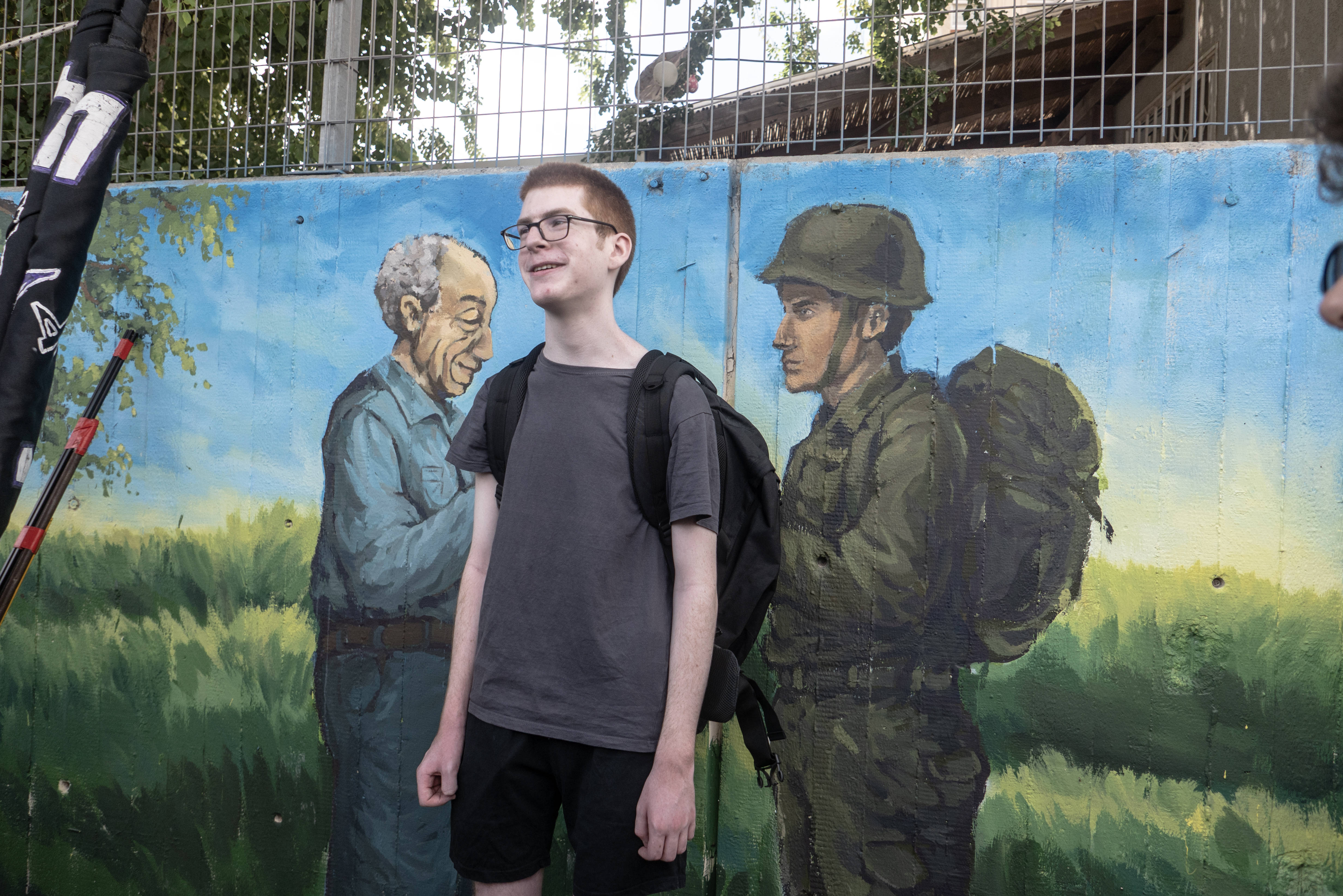 Ein junger Mann vor einer Mauer, dort zu sehen, ein Graffiti mit einem alten Mann und einem Soldaten