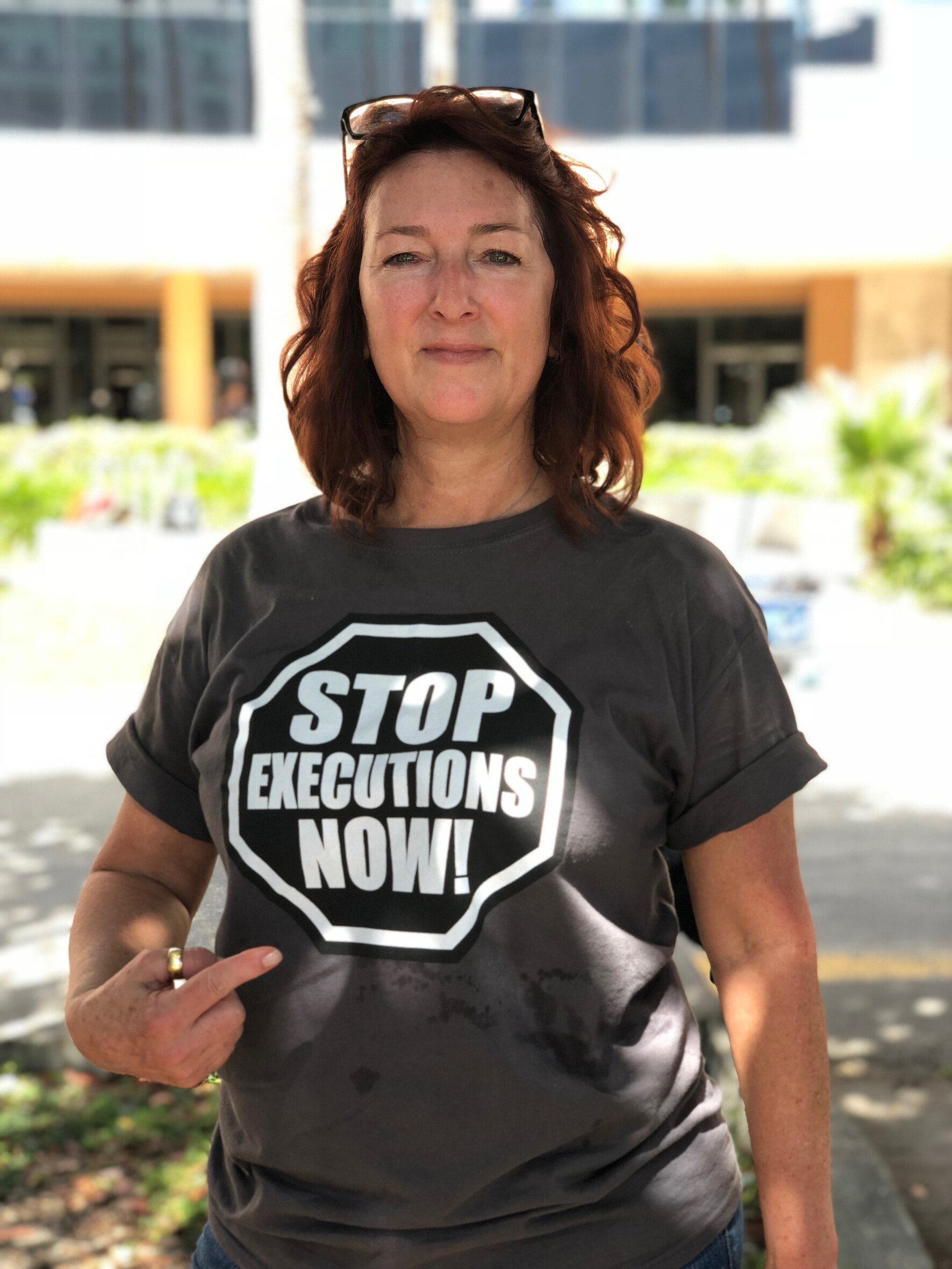 Aufnahme einer Frau an einem sonnigen Tag. Sie blickt leicht lächelnd in die Kamera und zeigt mit ihrer rechten Hand auf ihr T-Shirt, auf dem grop steht: "Stob executions now!"