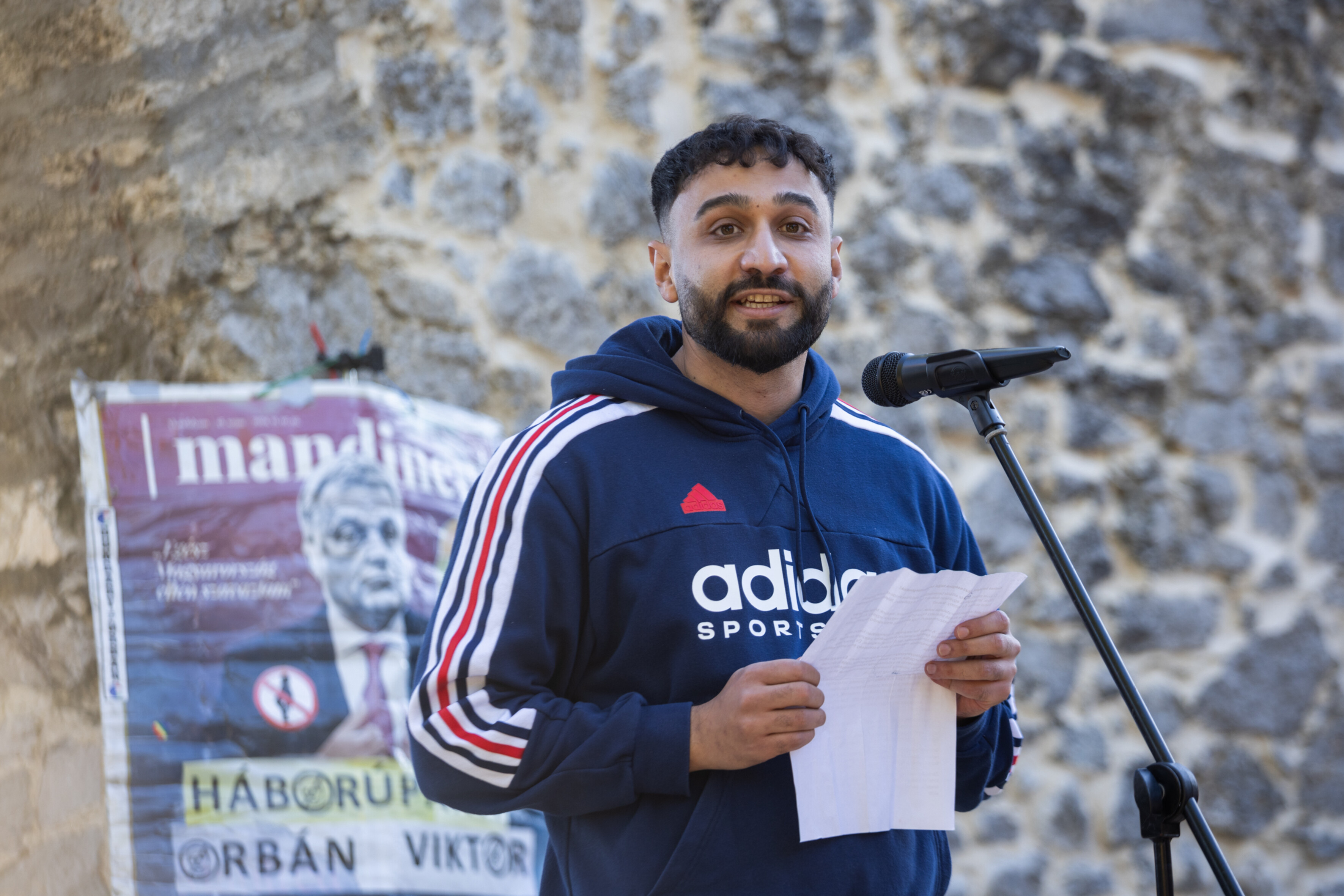 Géza Buzás-Hábel hält eine Rede. Vor ihm steht ein Mikrofon. Er trägt einen Kapuzenpullover. An einer Mauer hinter ihm hängt ein Protestplakat mit dem Foto von Ungarns Ministerpräsident Viktor Orbán.