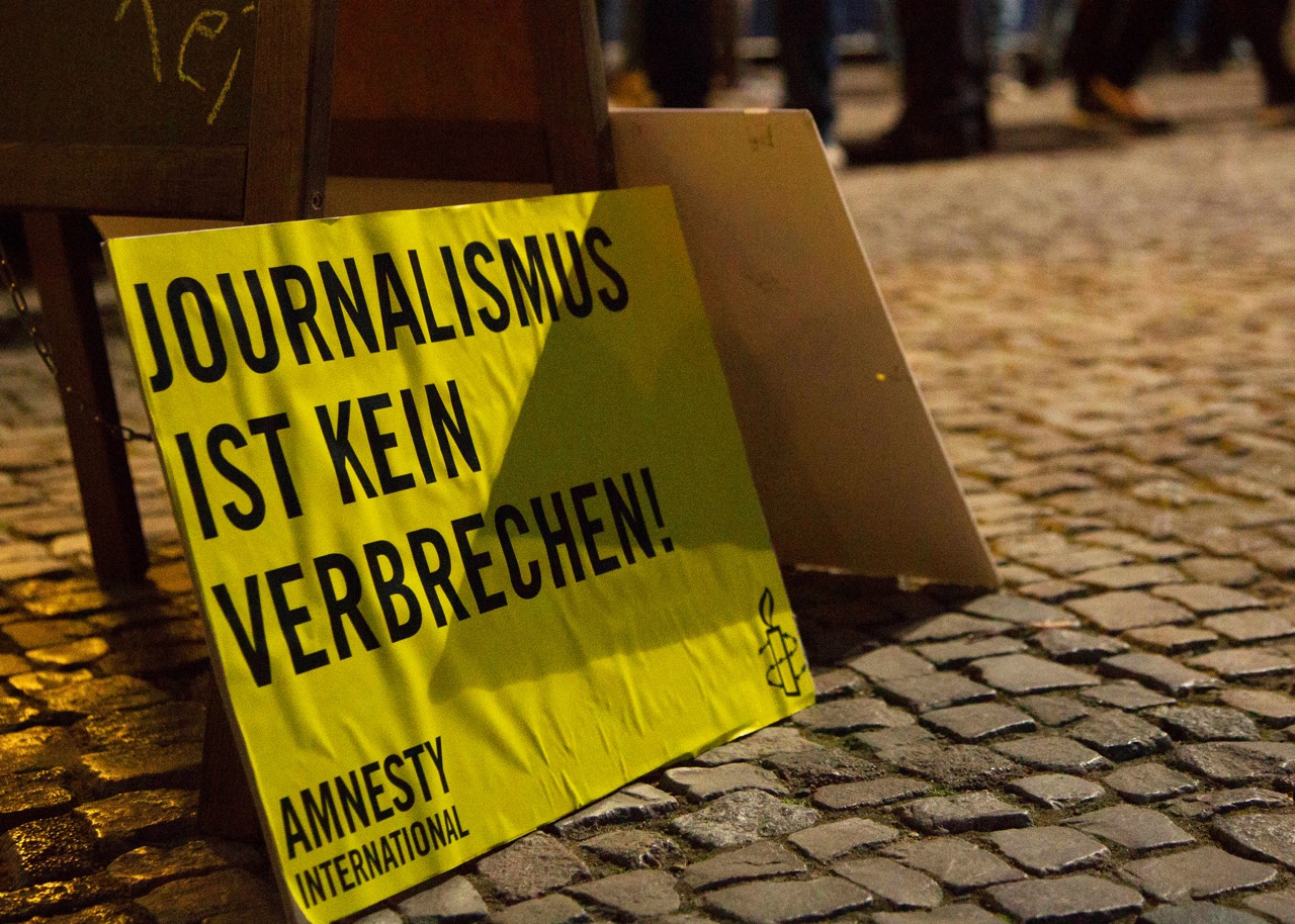 Das Foto zeigt ein Pappschild mit Amnesty-Logo und dem Schriftzug "Journalismus ist kein Verbrechen" auf dem Kopftseinpflaster eines Platzes stehend.