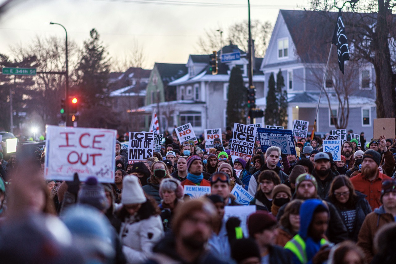 Protestug mit vielen Menschen zieht durch eine Straße. Die Menschen halten unter anderem Schilder hoch, auf denen steht "ICE out".