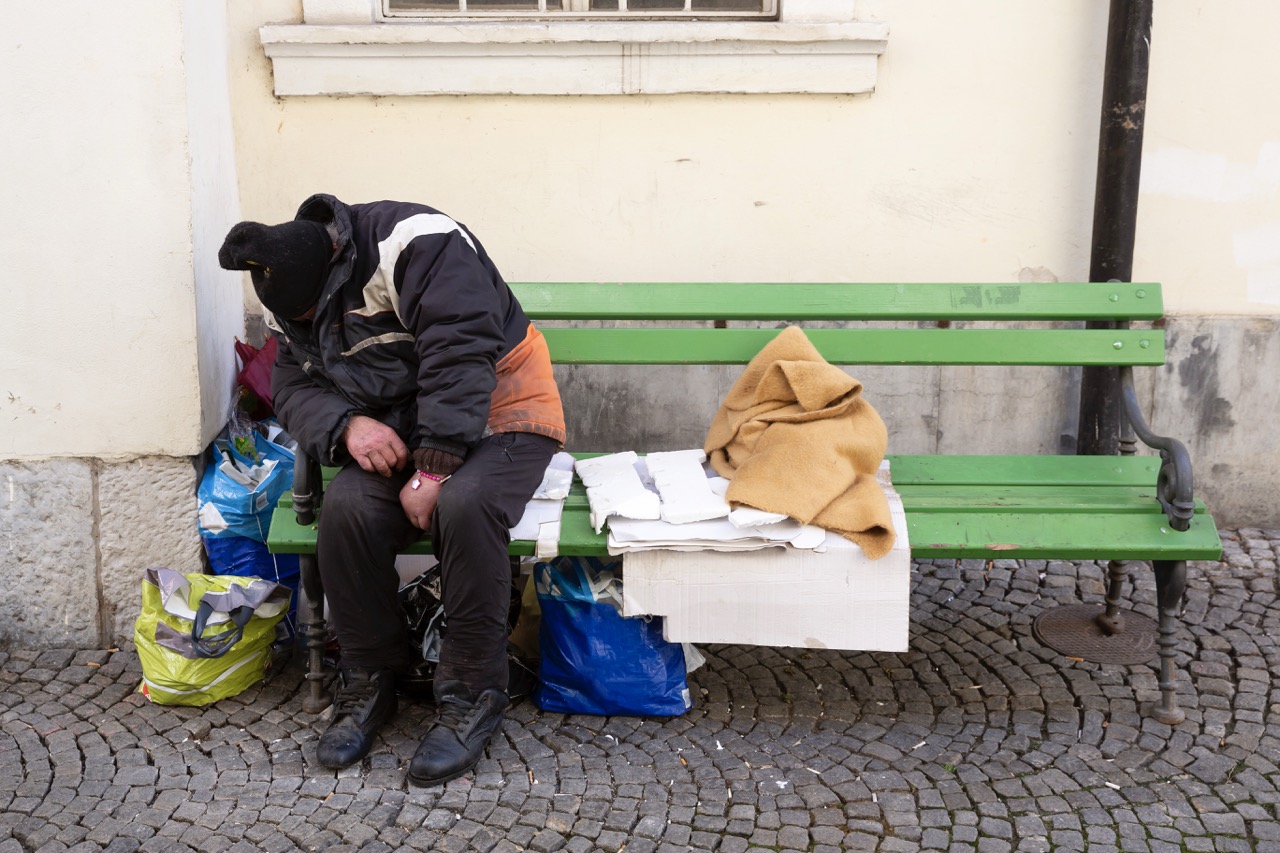 Ein Mann sitzt zusammengesunken auf einer Parkbank. Er scheint zu schlafen. Er trägt eine dicke Jacke und eine Mütze. Um ihn herum stehen Tüten mit seinen Habseligkeiten.