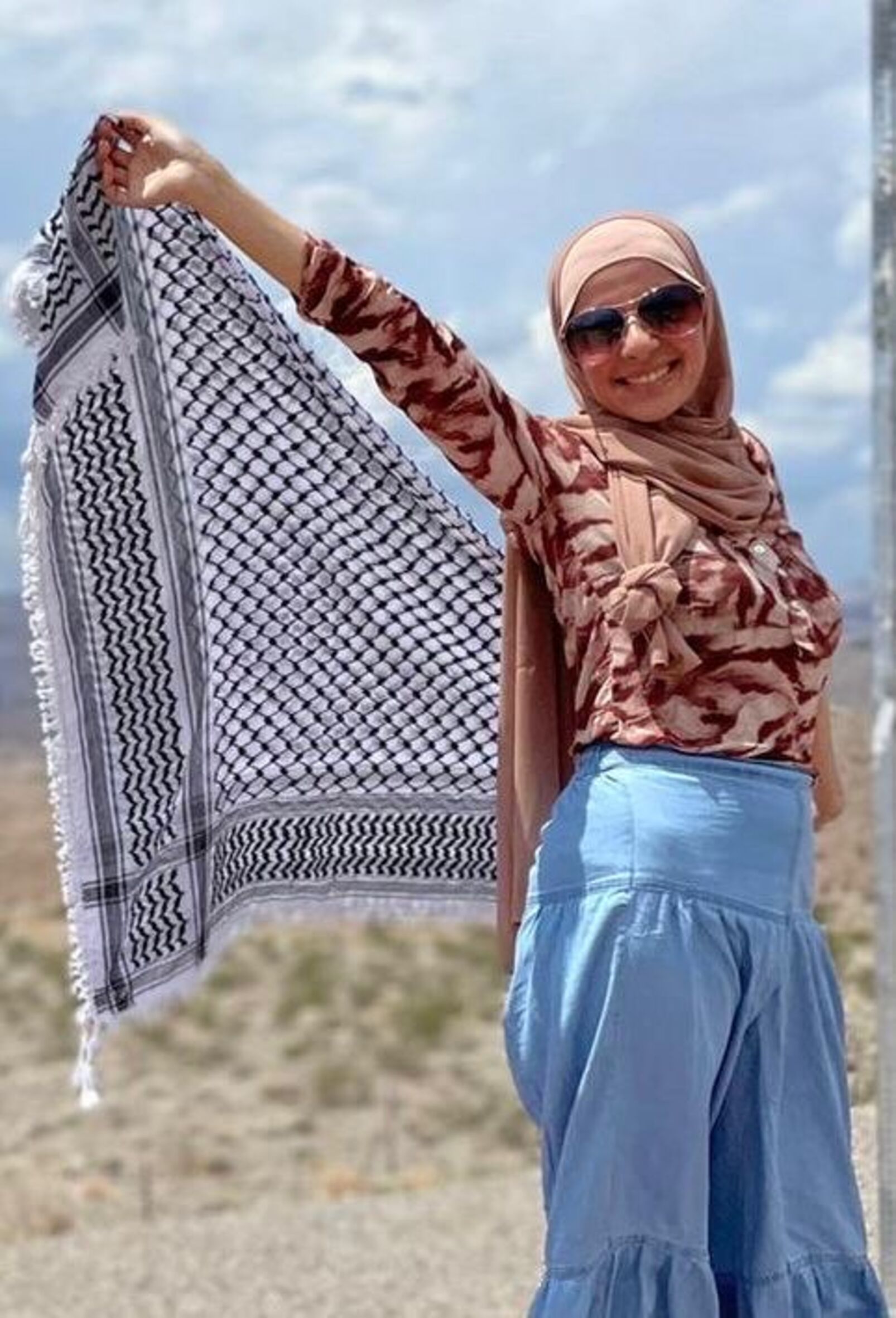 Das Foto zeigt Leqaa Kordia. Sie trägt ein Kopftuch und eine Sonnebrille und lächelt in die Kamera. Sie breitet mit ihren Händen hinter ihrem Rücken ein großes Kufiya-Tuch aus.