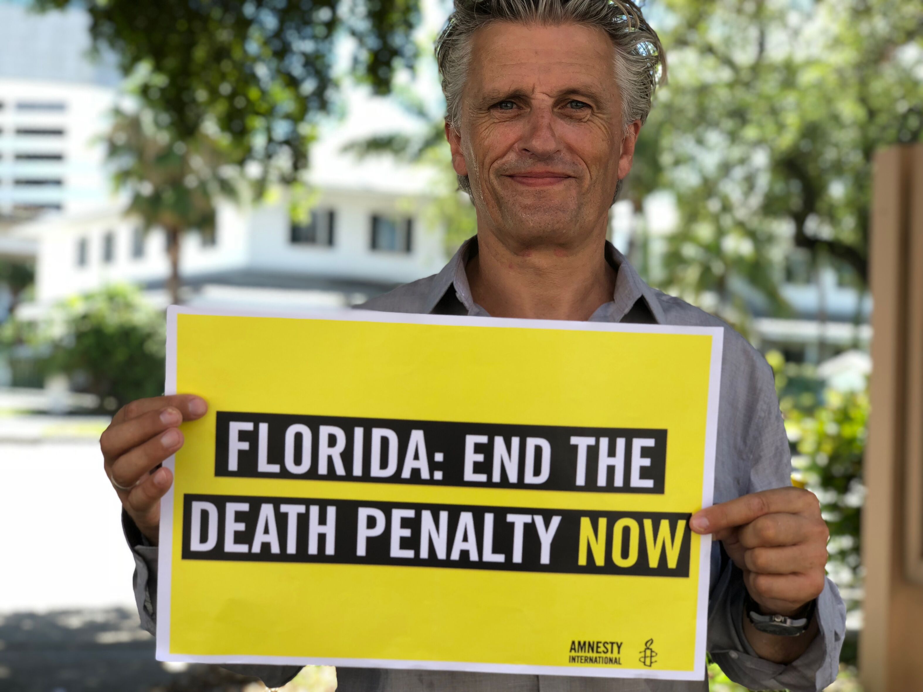 Aufnahme eines Mannes an einem sonnigen Tag. Er blickt leicht lächelnd in die Kamera und hält ein Schild mit Amnesty-Logo vor sich. Auf dem Schild steht: "Florida: End the Death Penalty now"