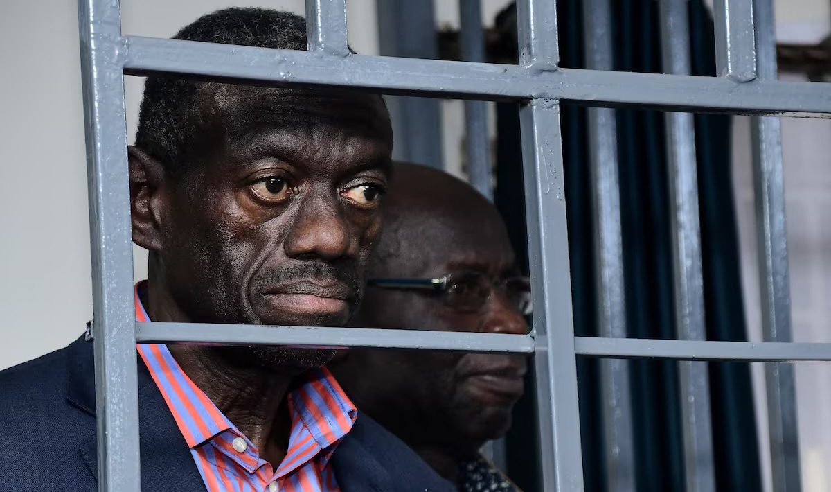 Der ugandische Oppositionspolitiker Dr. Kizza Besigye in einem dunkelblauen Sakko und orange-blau gestreiften Hemd hinter grauen Gefängnisstäben.
