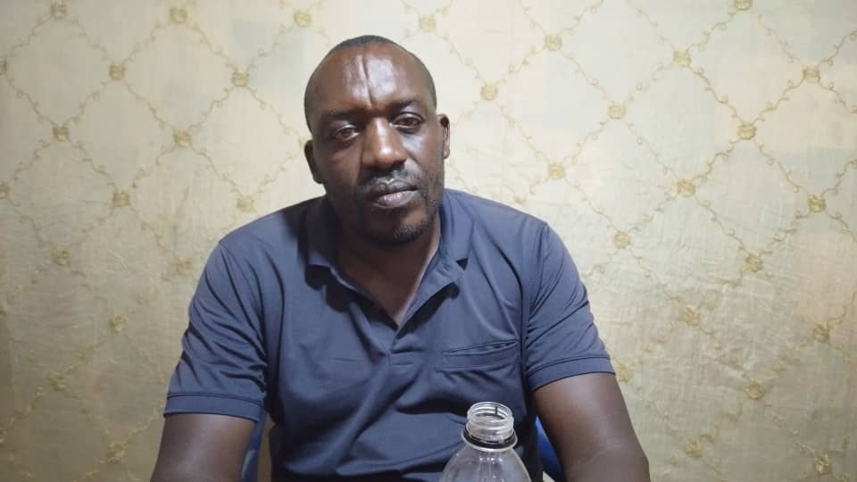 Das Foto zeigt Athanase Mvuyekure in einem Zimmer sitzen. Er hält eine Plastikflasche in der Hand und schaut ernst in die Kamera.