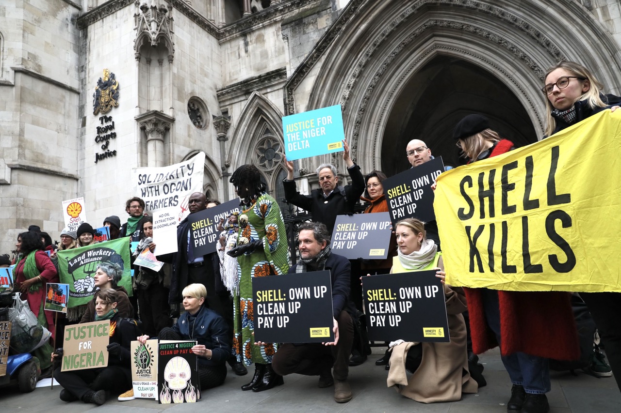 Circa 15 Personen stehen und hocken dich nebeneinander vor einem Gebäuden. Sie halten Schilder und Plakate hoch, auf denen unter anderem steht "Justice for the Niger Delta" oder "Shell kills".