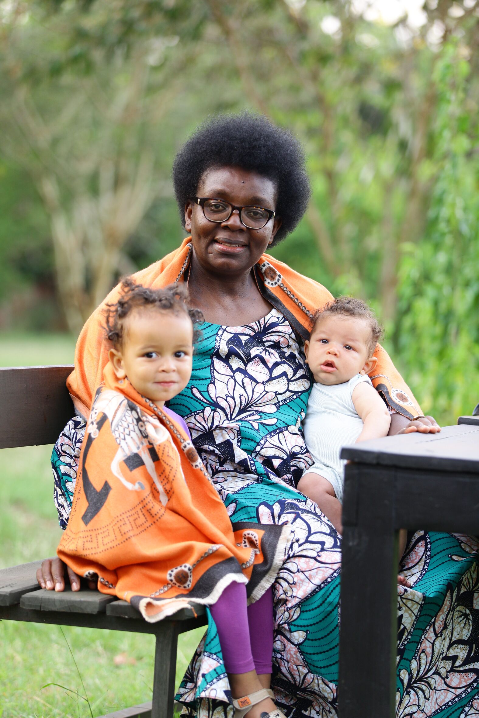 Das Foto zeigt Victoire Ingabire auf einer Bank im Freien sitzen auf einer Wiese. Hinter ihr stehen Bäume und Büsche. Rechts und links neben ihr sitzt jeweils ein kleines Kind. Alle lächeln.