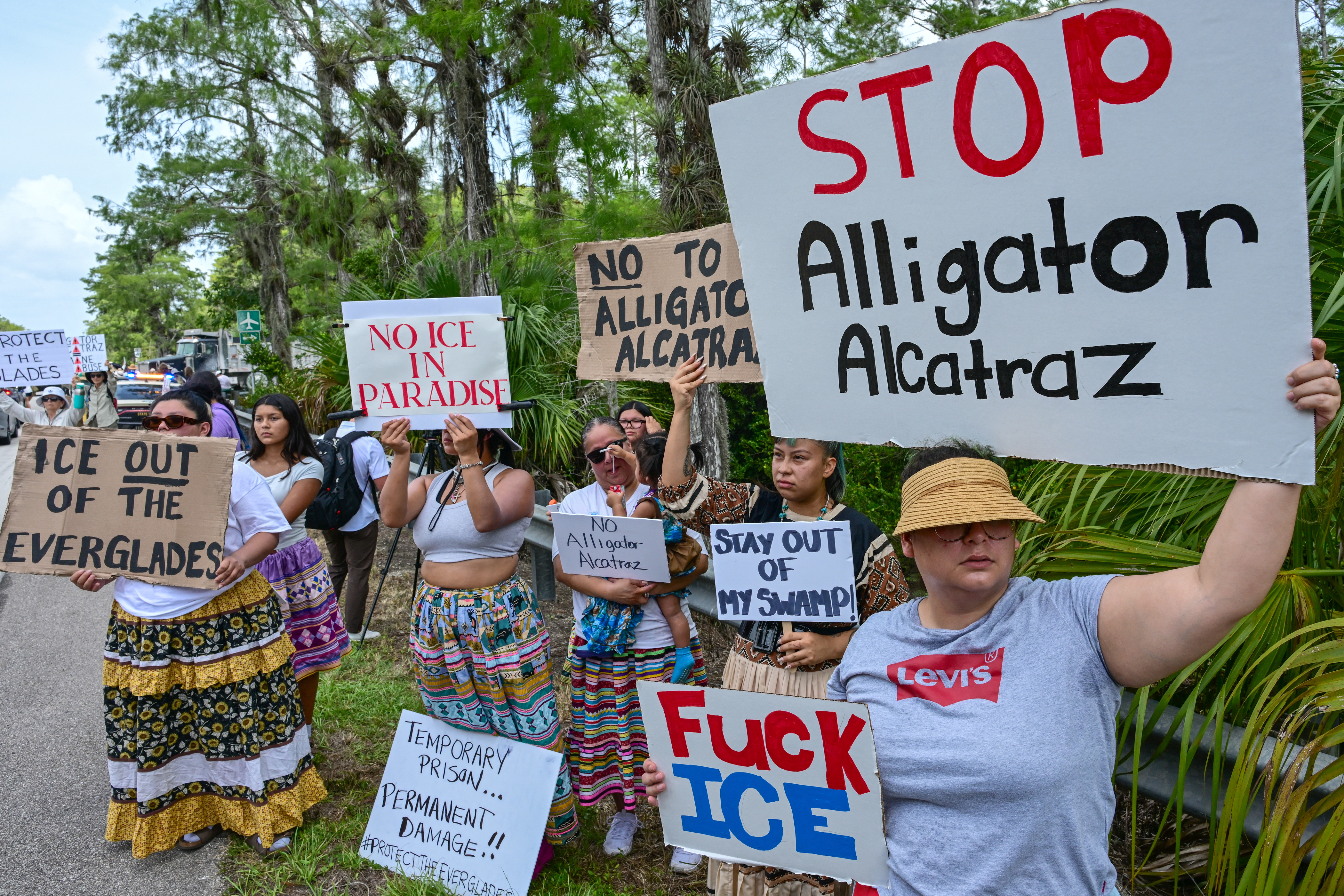 Eine Gruppe von Demonstranten hält am Rande eines Naturgebiets Plakate mit Parolen wie "STOP Alligator Alcatraz" und "ICE OUT OF THE EVERGLADES" in die Kamera. Sie protestieren damit sichtbar gegen die US-Einwanderungsbehörde ICE und ein geplantes Bauprojekt.