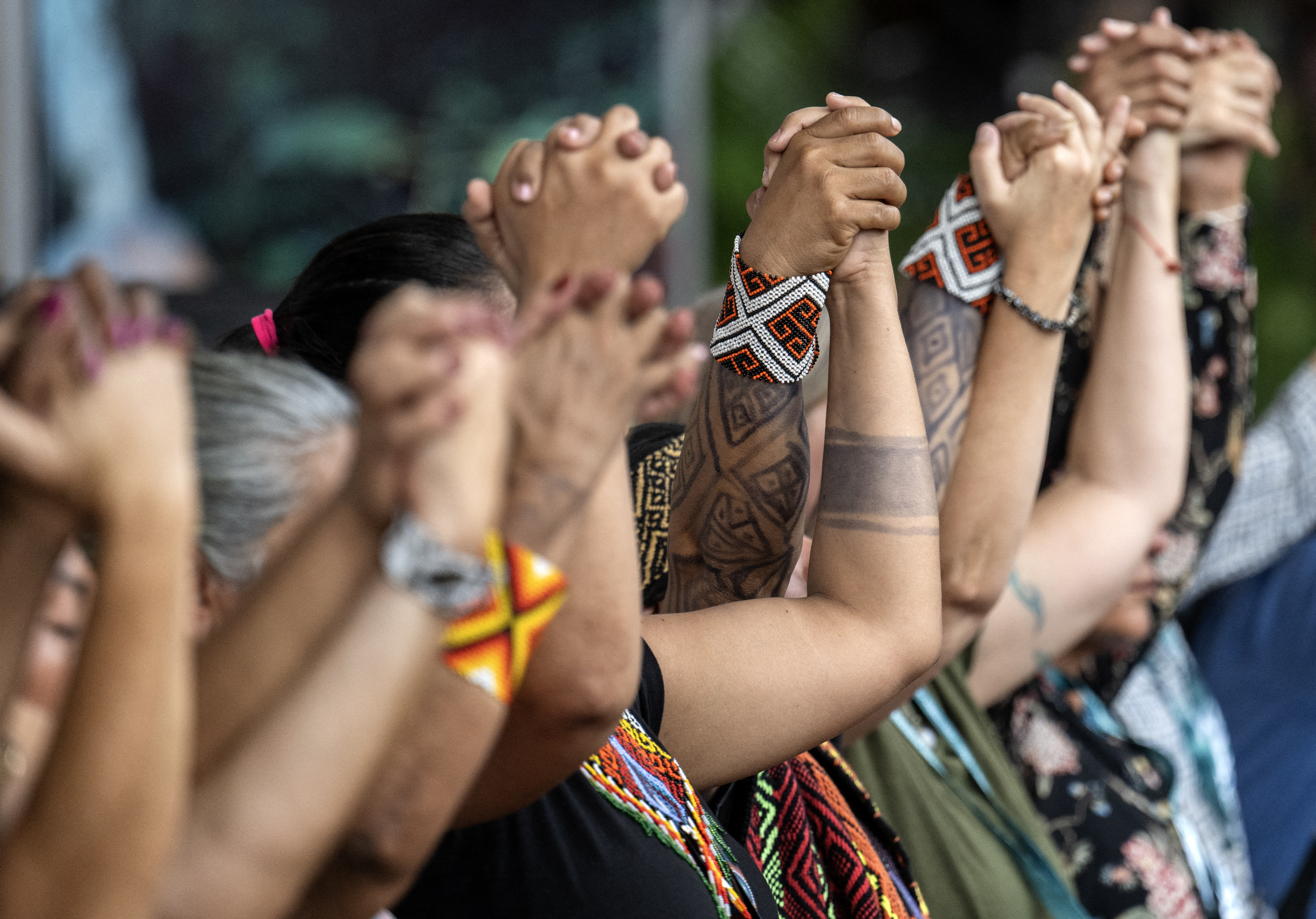 Eine Gruppe von Menschen hält ihre ineinander verschränkten Hände gemeinsam nach oben gerichtet. An den Handgelenken einiger Personen sind auffällige, bunt gewebte Armbänder mit geometrischen Mustern zu erkennen.