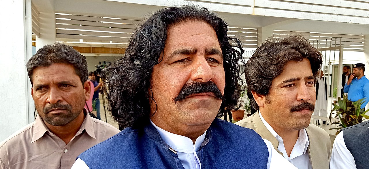 Nahaufnahme von Ali Wazir. Er trägt einen Schnauzbart und blickt ernst. Links und rechts von ihm steht jeweils ein Mann.
