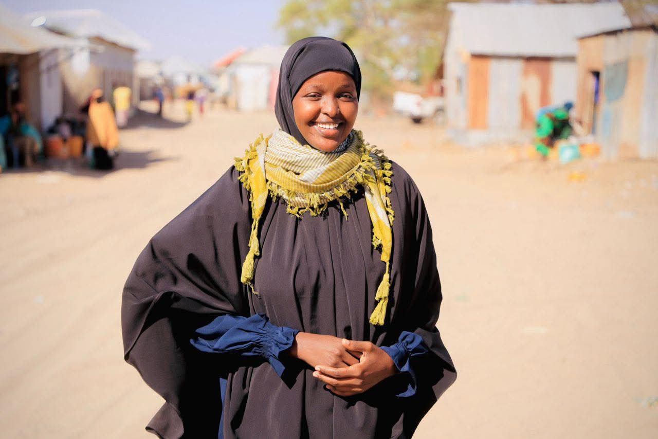 Das Foto zeigt Sadia Moalim Ali auf einer nicht asphaltierten Straße. Sie trägt ein Kopftuch und ein Gewand und lächelt in die Kamera. Im Hintergrund stehen einstöckige Gebäude.