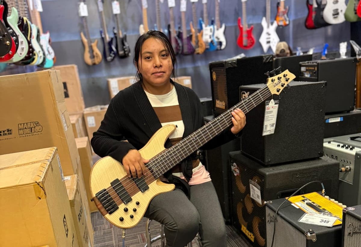 Das Foto zeigt Andrea Pedro-Francisco in einem Musikladen mit einem E-Bass in den Händen. Sie blickt in die Kamera und lächelt leicht. Hinter ihr hängen Gitarren an der Wand.
