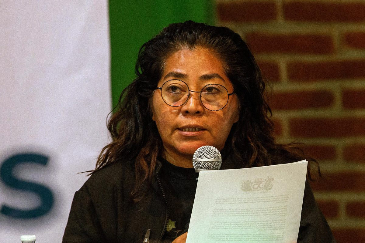 Das Foto zeigt Dora Roblero an einem Tisch sitzend. Sie spricht in ein Mikrofon und hält in der linken Hand einen Zettel. Sie trägt eine Brille.
