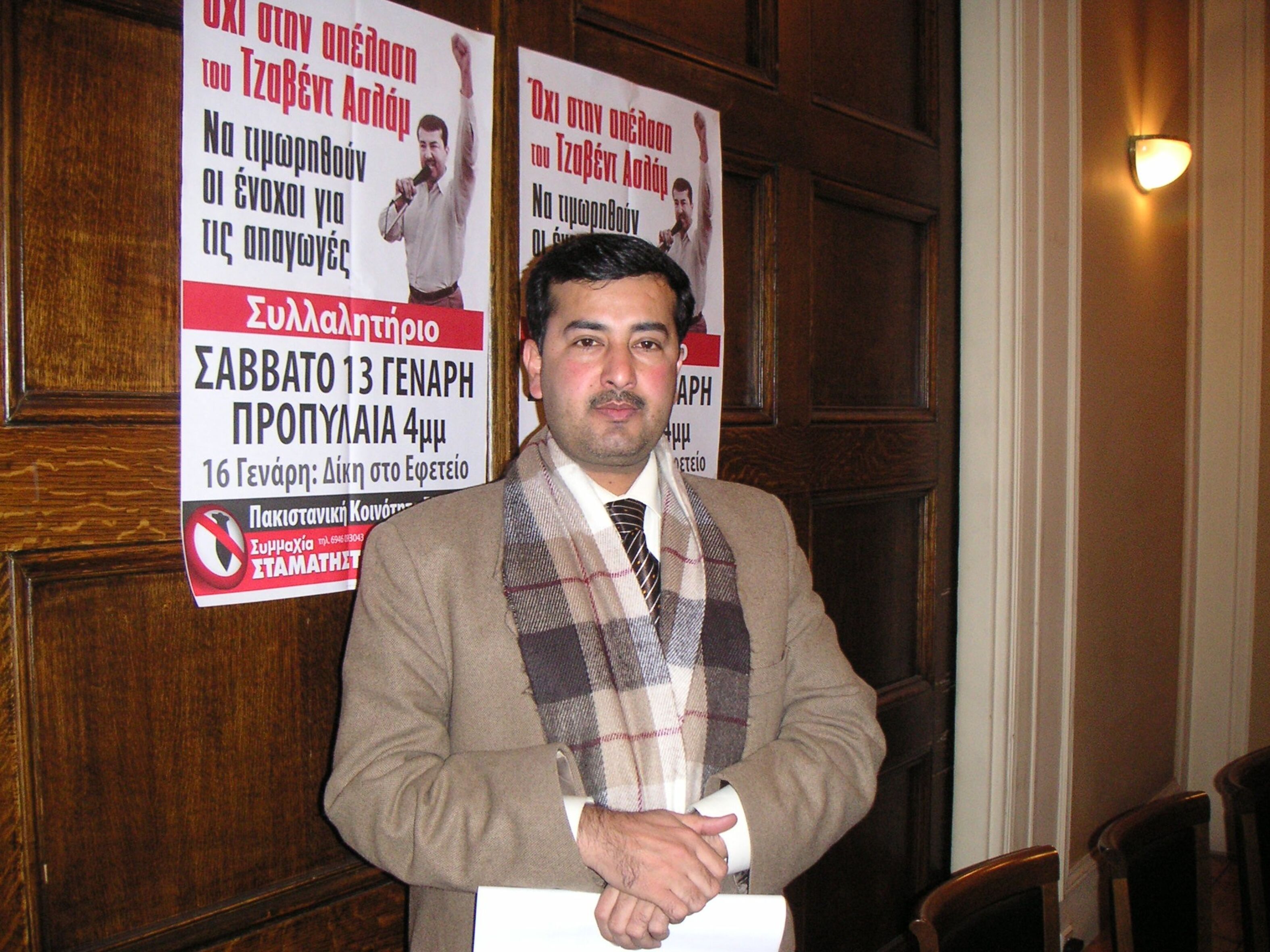 Foto von Javed Aslam, der Jacke, Schal und Krawatte trägt und in seinen gefalteten Händen ein Blatt Papier vor sich hält. Er steht vor einer holzvertäfelten Wand, auf der zwei Poster angebracht sind mit einem Mann, der seine linke Faust in die Höhe streckt.