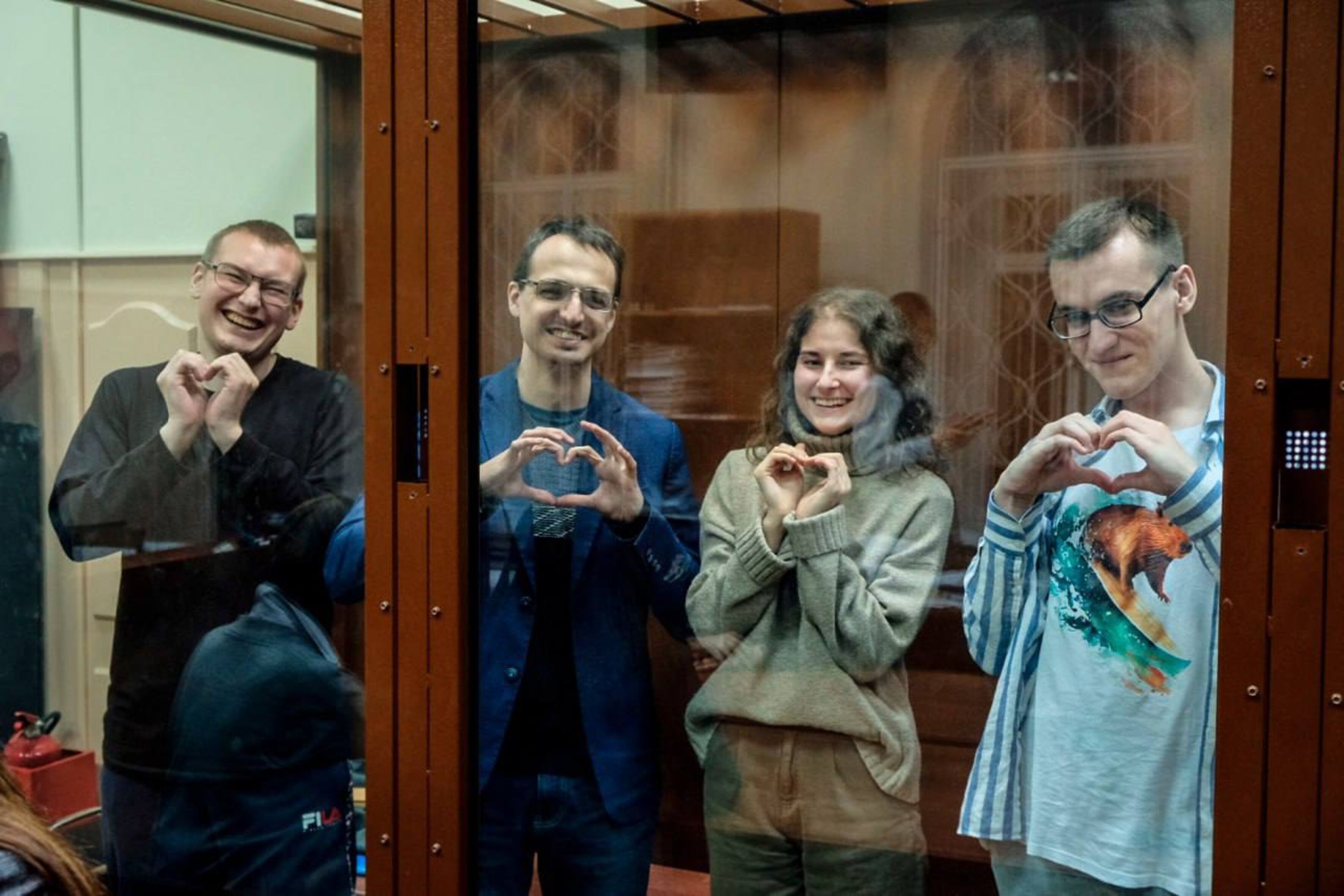 Das Foto zeigt die vier jungen Personen in einem Glaskasten. Ihre Hände sind mit Handschellen gefesselt. Sie formen ihre Hände zu einem Herzen und lachen in die Kamera. 
