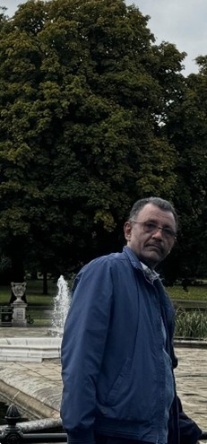 Foto von Mohammed Farouk Salman Mahmoud, aufgenommen in einem Park. Im Hintergrund stehen ein kleiner Springbrunnen und Bäume. Er trägt eine Jacke und eine Brille und blickt ernst in die Kamera.