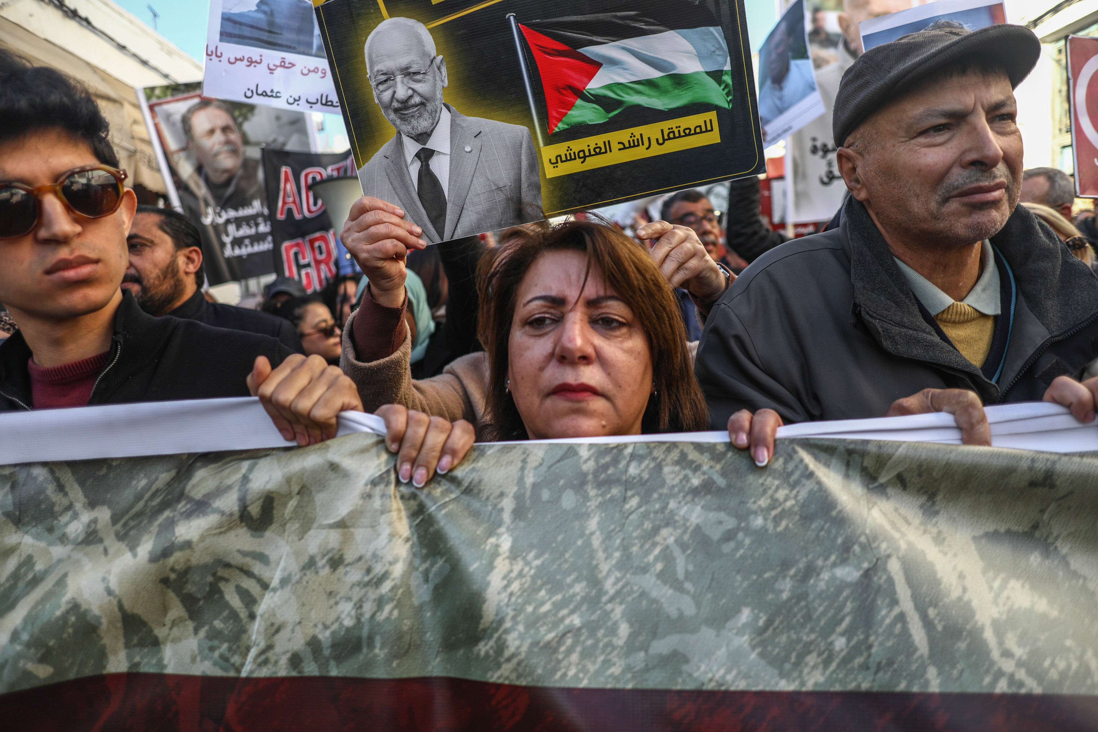 Das Foto zeigt einen jüngeren Mann, eine Frau und einen Mann, die bei einer Demonstration ein Banner vor sich hertragen. Hinter ihnen hält eine Person ein Plakt hoch mit dem Fotos Mannes.