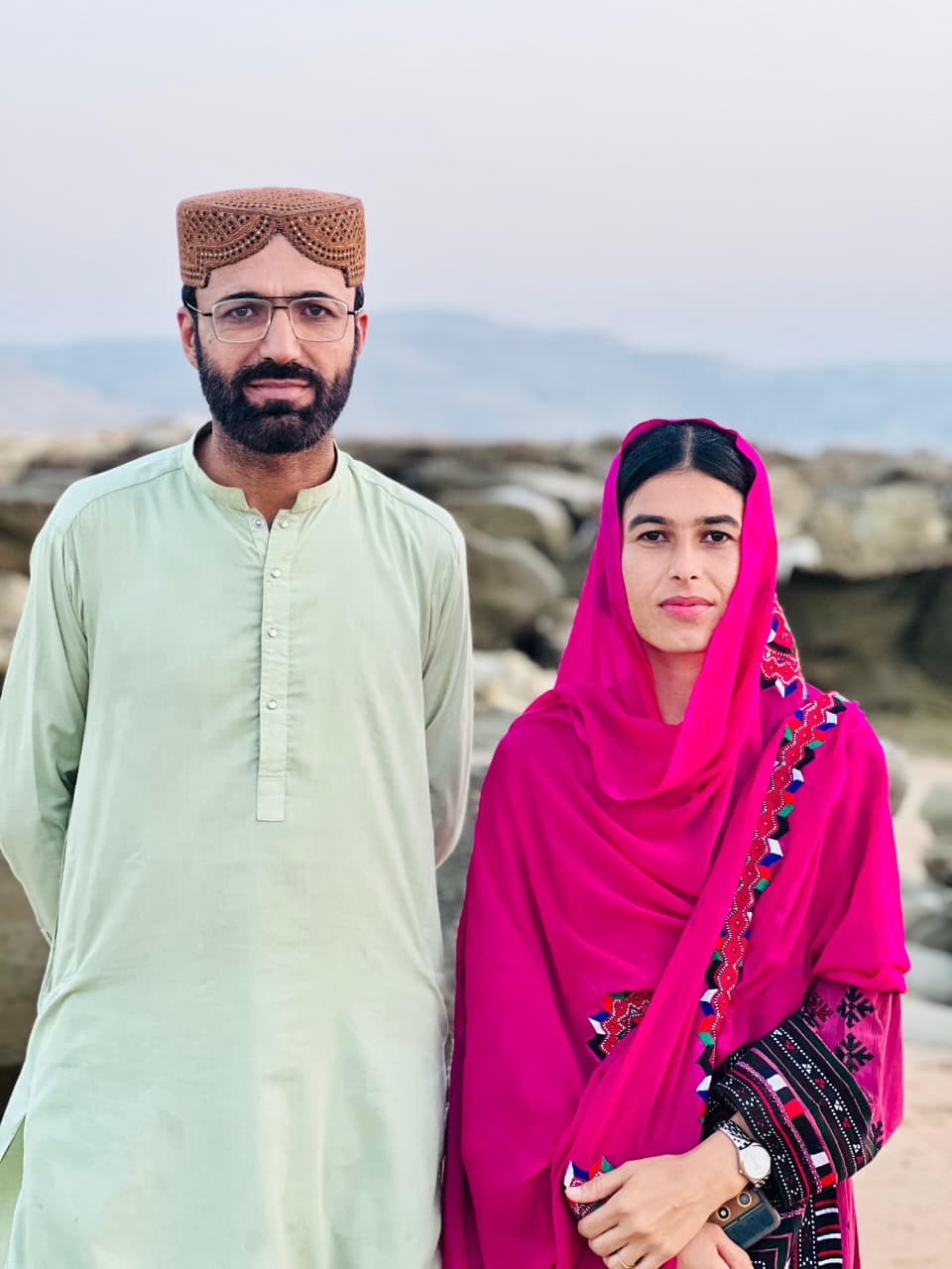 Das Foto zeigt Dad Shah und Fozia Baloc nebeneinander in einer Landschaft stehen und leicht in die Kamera lächelnd. Sie tragen traditionelle Kleidung.
