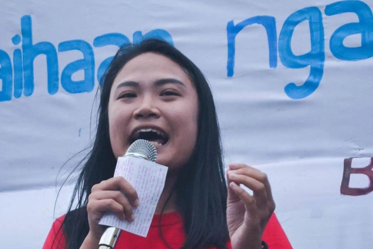 Das Foto zeigt Marielle Domequil, die auf einer Kundgebung eine Rede hält. In ihrer rechten Hand hält sie ein Mikrofon und einen Zettel. Hinter ihr sind zwei Banner angebracht.