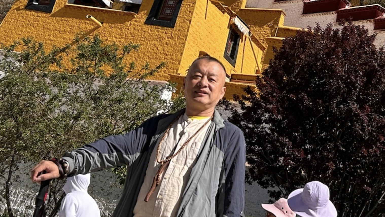 Das Foto zeigt Zhang Yi vor einem Gebäude stehen. Er blickt in die Kamera und hat seine rechte Hand auf einer Art Wanderstock gestützt.