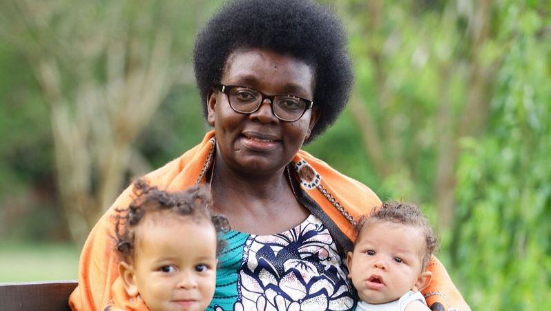 Das Foto zeigt Victoire Ingabire auf einer Bank im Freien sitzen auf einer Wiese. Hinter ihr stehen Bäume und Büsche. Rechts und links neben ihr sitzt jeweils ein kleines Kind. Alle lächeln.