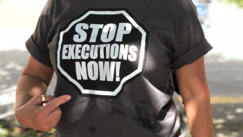 Aufnahme einer Person an einem sonnigen Tag, die mit ihrer rechten Hand auf ihr T-Shirt zeigt, auf dem steht: "Stob executions now!"