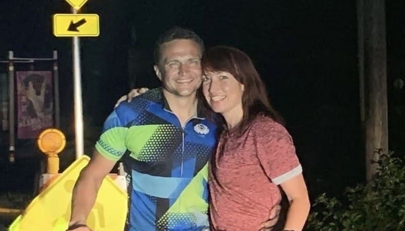 Das Foto zeigt Andriy Shepitsen, der in Sportkleidung hinter einem Fahrrad steht und in die Kamera lächelt. Neben ihm steht eine Frau, die ebenfalls lächelt. Sie umarmen sich gegenseitig. Das Foto wurde abends oder nachts aufgenommen vor einem Verkehrsschild.