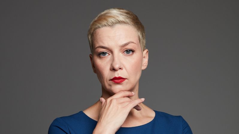 Porträtaufnahme von Maryia Kalesnikava, die ein Kleid trägt und kurze blonde Haare und ernst in die Kamera schaut, während sie ihre recht Hand unter ihr Kinn hält.