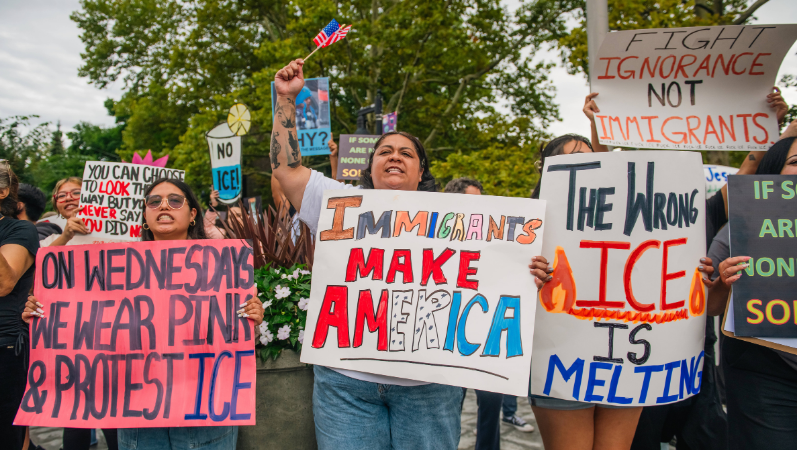 Das Foto zeigt mehrere Frauen bei einer Demonstration. Sie halten Schilder hoch. Auf einem steht: "The wrong ICE is melting". Auf einem anderen: "Immigrants make America".