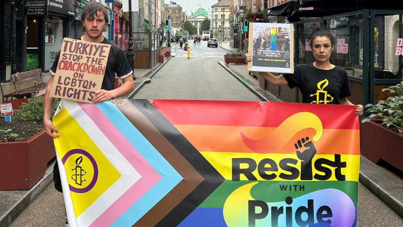 Das Foto zeigt einen jungen Mann und eine junge Frau in einer schmalen Straße. Sie tragen Amnesty-T-Shirts und halten die Intersex-Inclusive-Pride-Flagge vor sich, auf dem steht "Resist with pride". Sie halten außerdem kleine Plakate in der Hand. Auf dem des Mannes steht: "Turkiye, stop the crackdown on LGTBIQ+ rights".