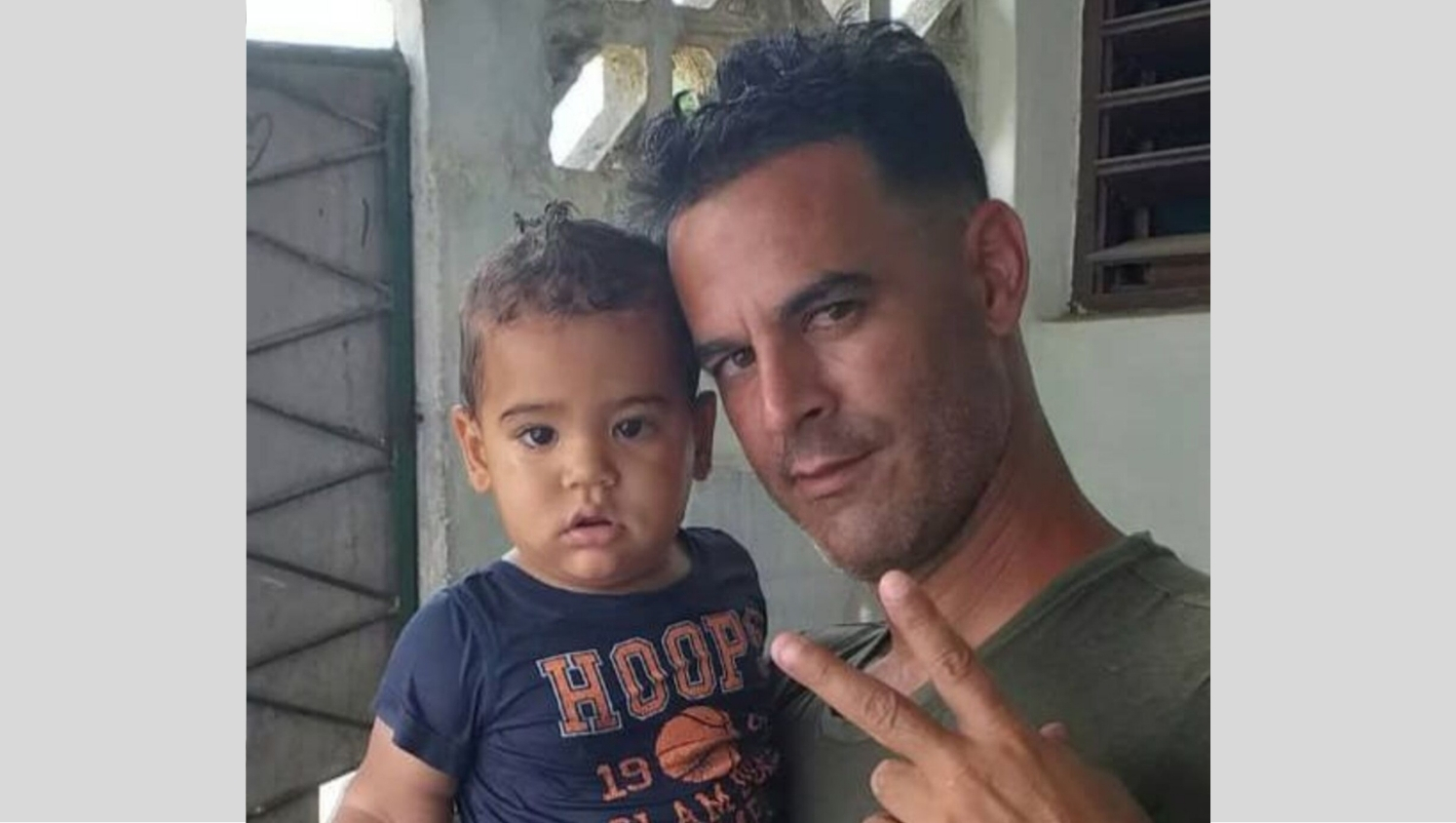 Roberto Pérez Fonseca hält ein kleines Kind in seinem rechten Arm. Beide blicken in die Kamera. Fonseca hält den den Zeige- und Mittelfinger seiner linken Hand gespreizt nach oben.