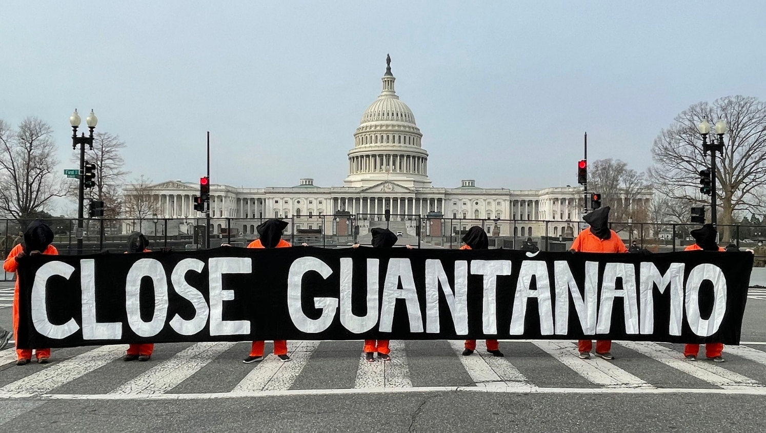 Mehrere Personen in Gefangenenkleidung und mit Säcken über den Kopf halten auf einer Straße ein großes Banner vor sich mit der Aufschrift: "Close Guantanamo". Im Hintergrund steht ein großes Geäude: Das Kapitol.