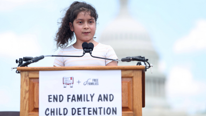 Ein Mächen steht an einem Renderpult und spricht in zwei Mikrofone. Auf dem Pult steht auf einem Plakat: "End family and child detention". Im Hintergund steht das Kapitol.