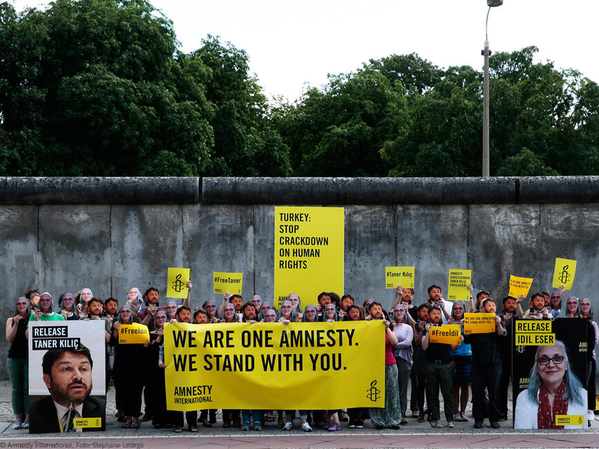 Eine Gruppe von Amnesty-Aktivistinnen und Aktivisten steht vor der Berliner Mauer, hält Banner und Schilder hoch, mit Masken von İdil Eser und Taner Kılıç